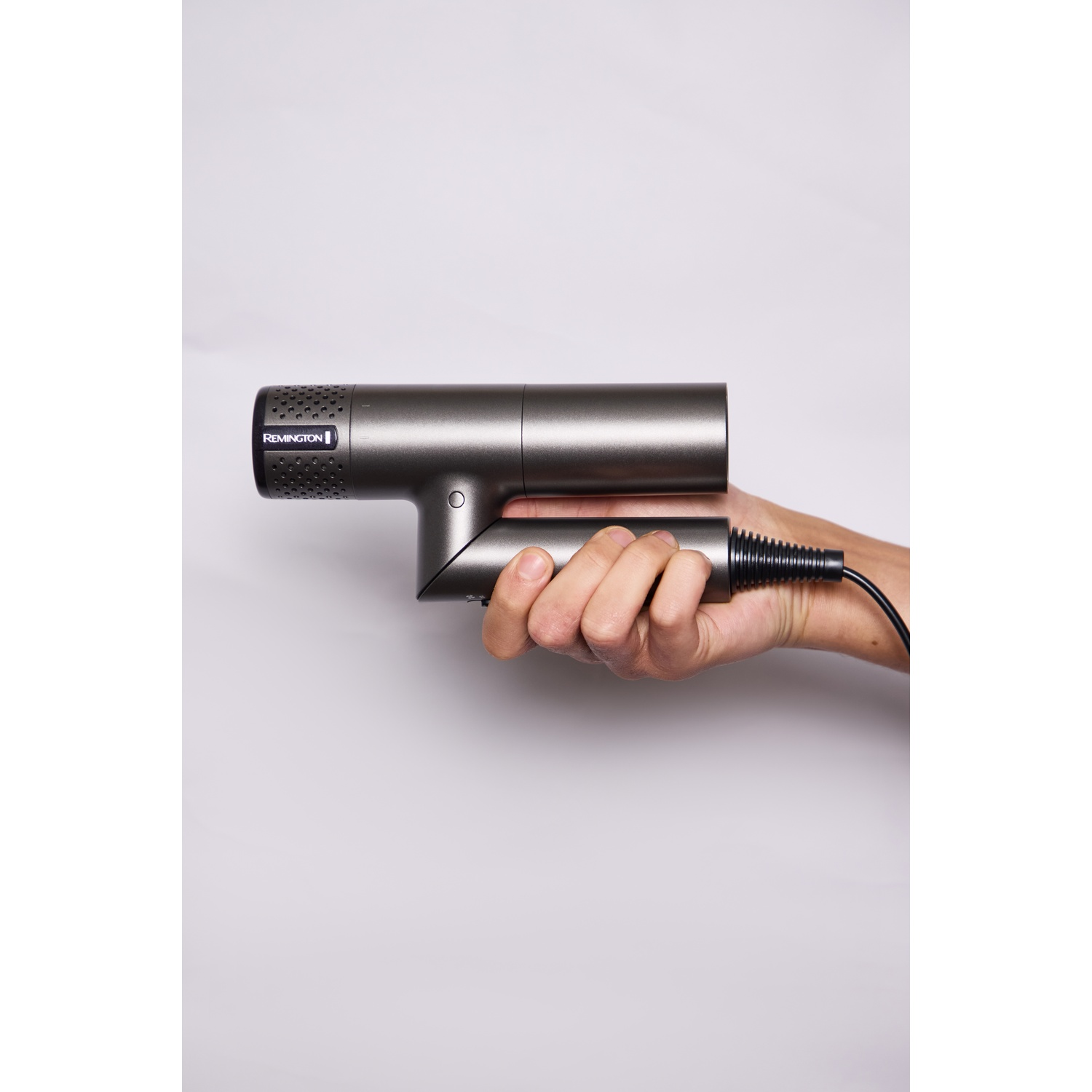 Een hand houdt een compacte, donkergrijze Remington föhn met opvouwbare handgreep voor een lichte achtergrond.