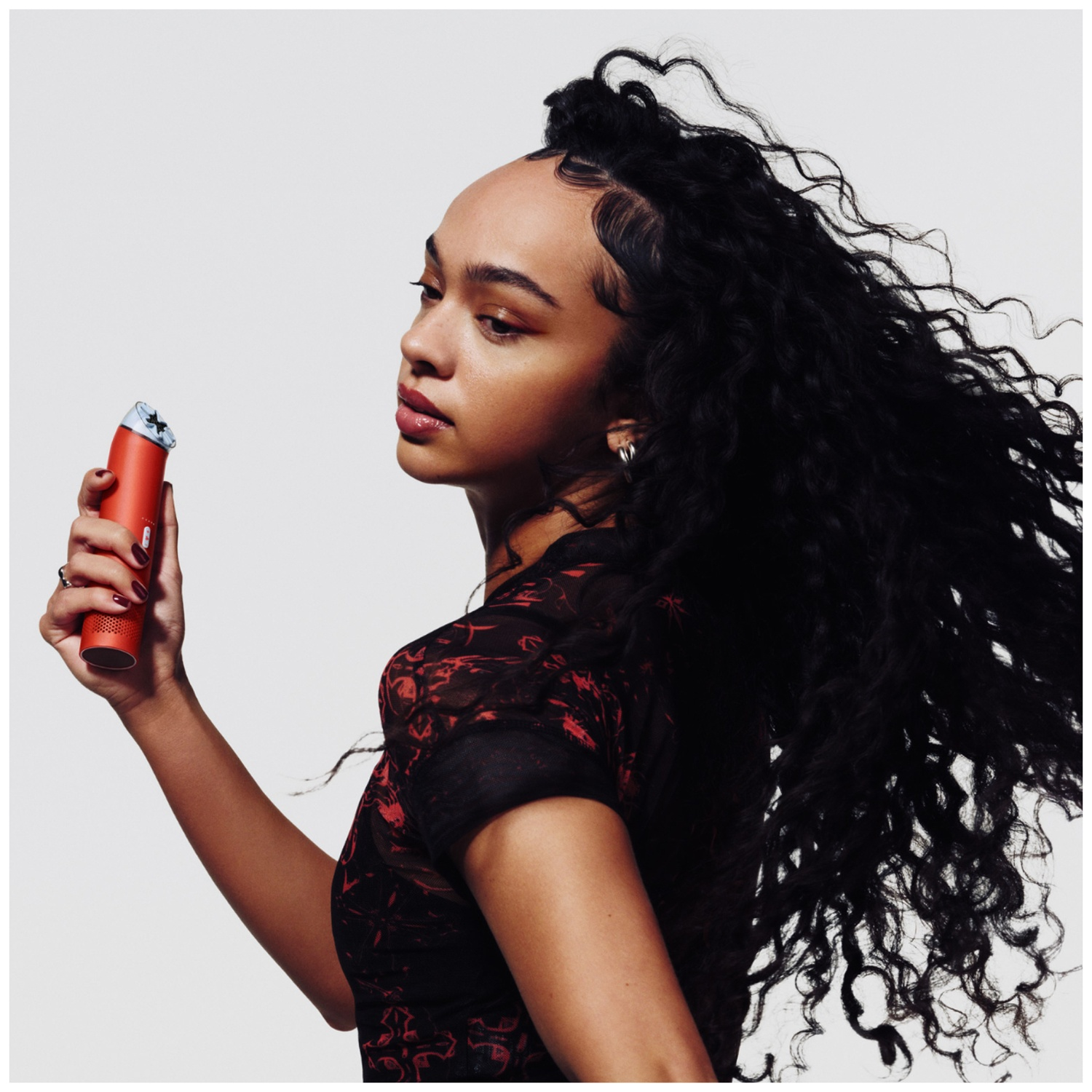 Une femme aux longs cheveux bouclés tient un objet rouge sur fond blanc.