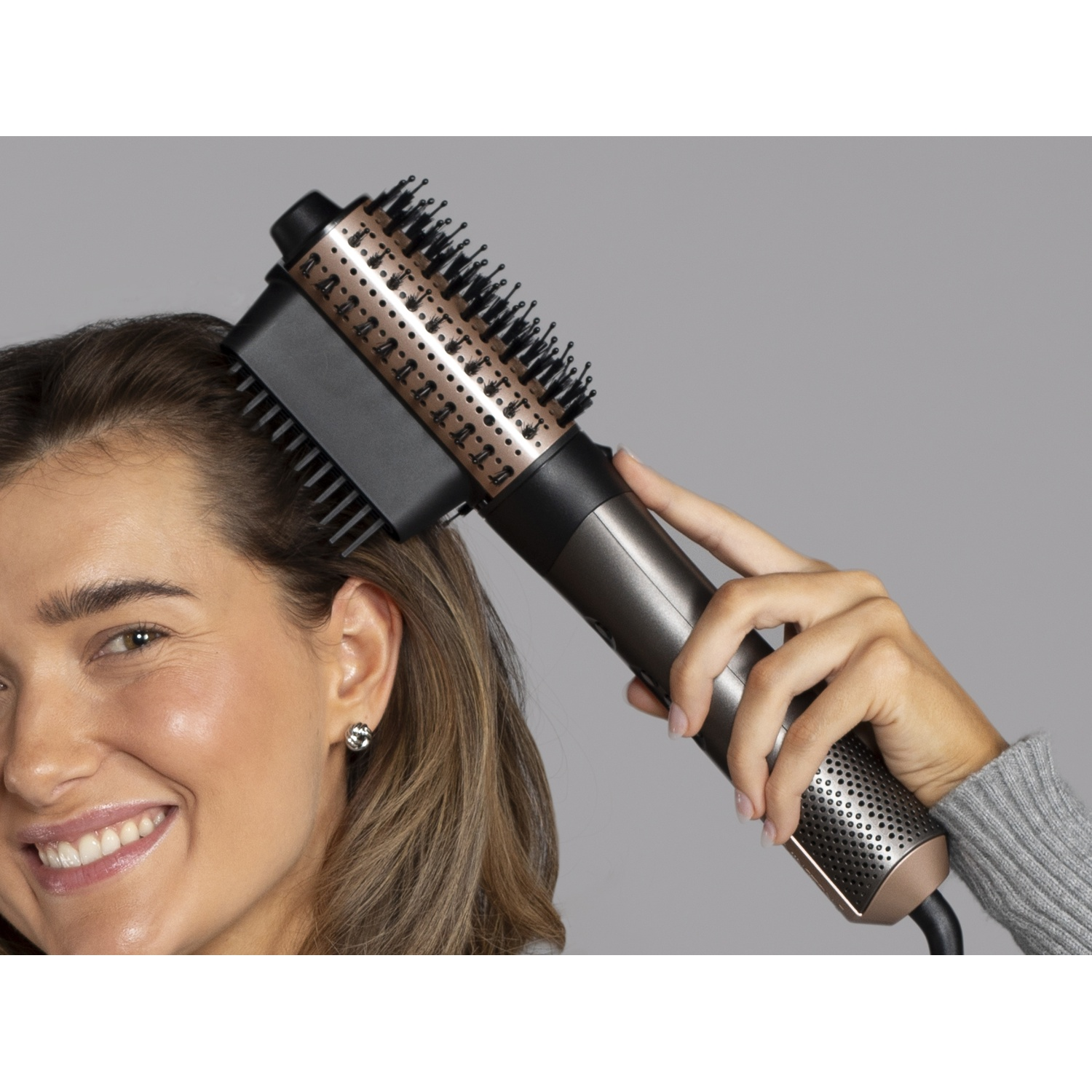 Une femme souriante coiffe ses cheveux bruns avec une brosse soufflante noire et dorée sur fond gris.