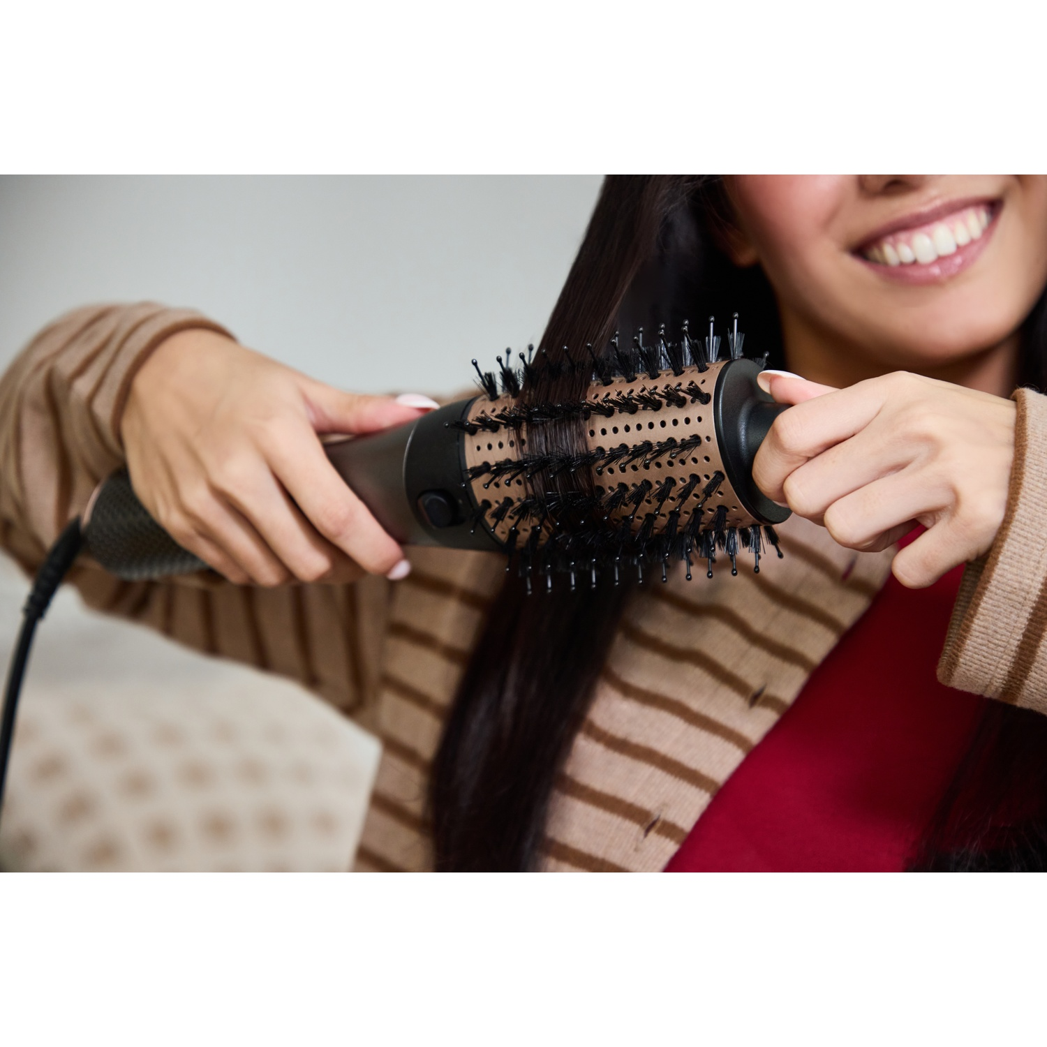 Une femme souriante utilise une brosse soufflante or rose pour coiffer ses longs cheveux sombres.
