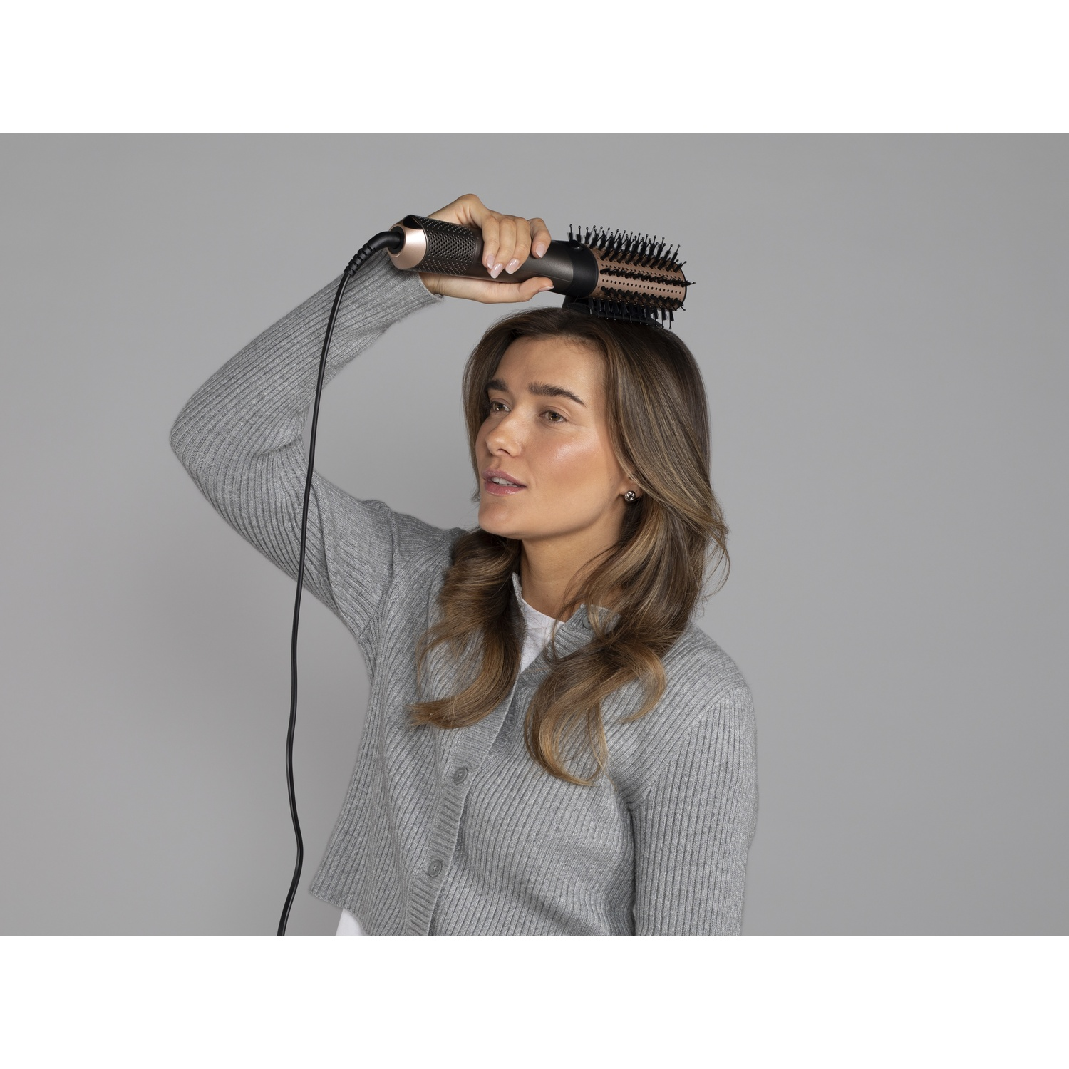 Une femme en gilet gris tient une brosse soufflante près de ses cheveux sur un fond gris.