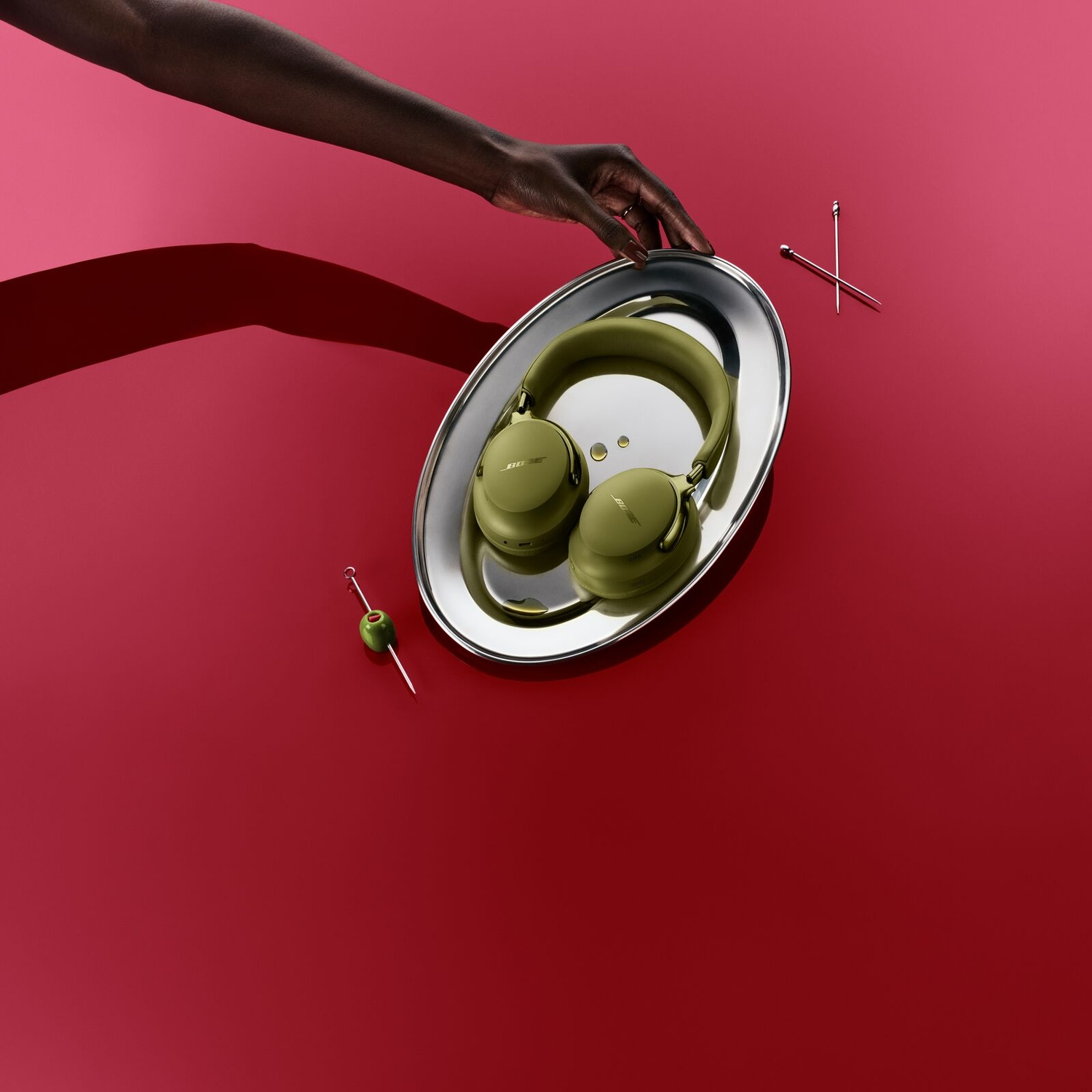 A hand places a silver tray containing green headphones and an olive on a red table.