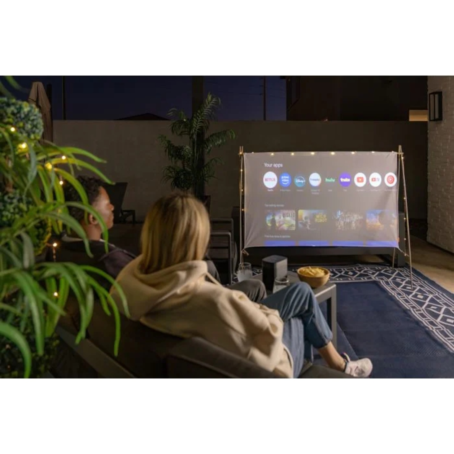 Un couple est assis sur un canapé extérieur regardant un écran de projecteur portable la nuit.