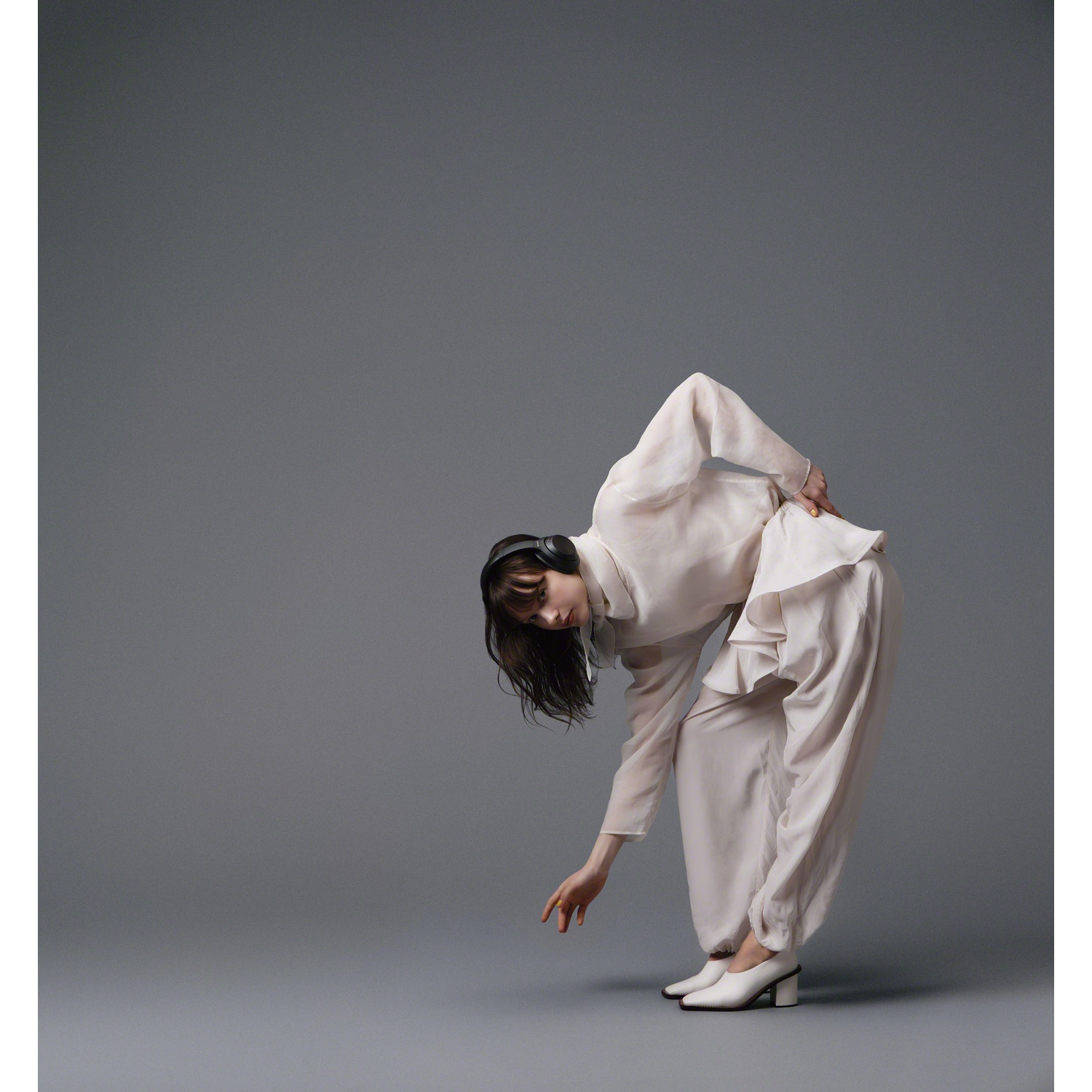 A woman in white clothing and headphones bends forward against a gray background.