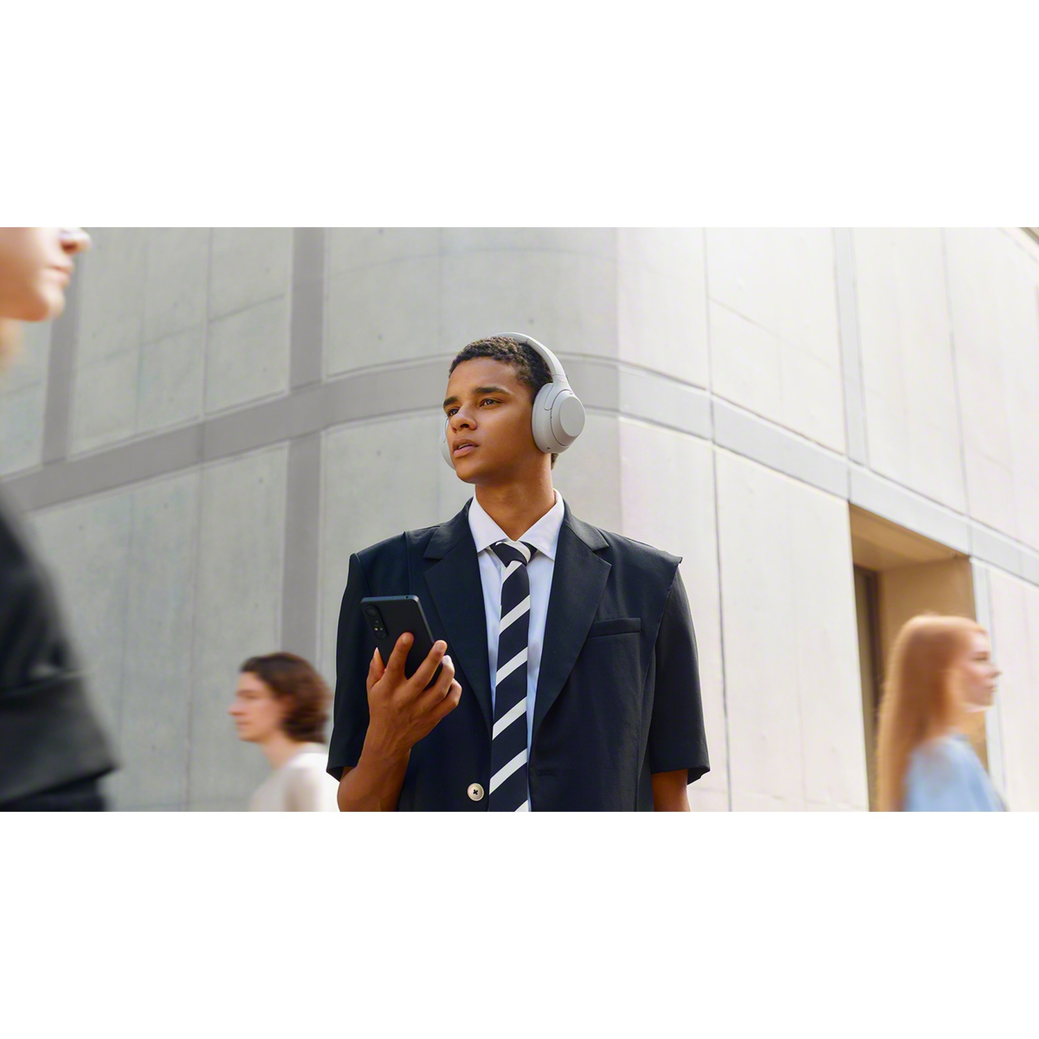 A young man in a suit wearing headphones and holding a smartphone stands on a busy street in front of a building.