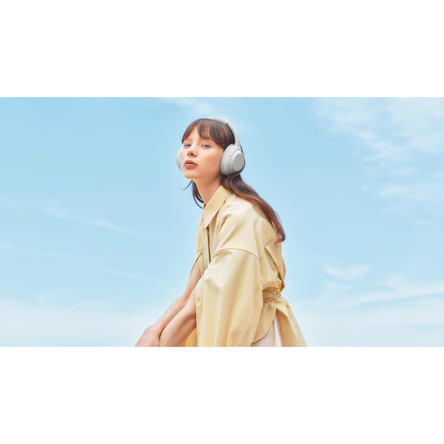 A young woman with brown hair and a beige coat wears gray headphones against a bright sky.