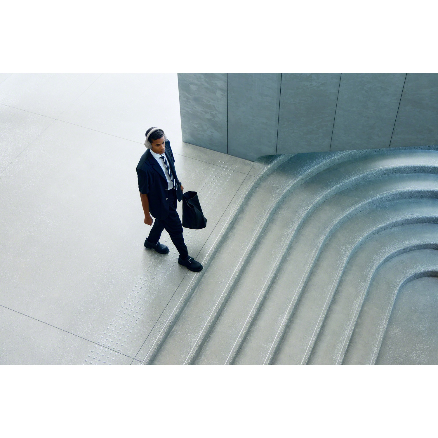 A man in a suit wearing headphones walks down stairs in a modern building, carrying a bag.