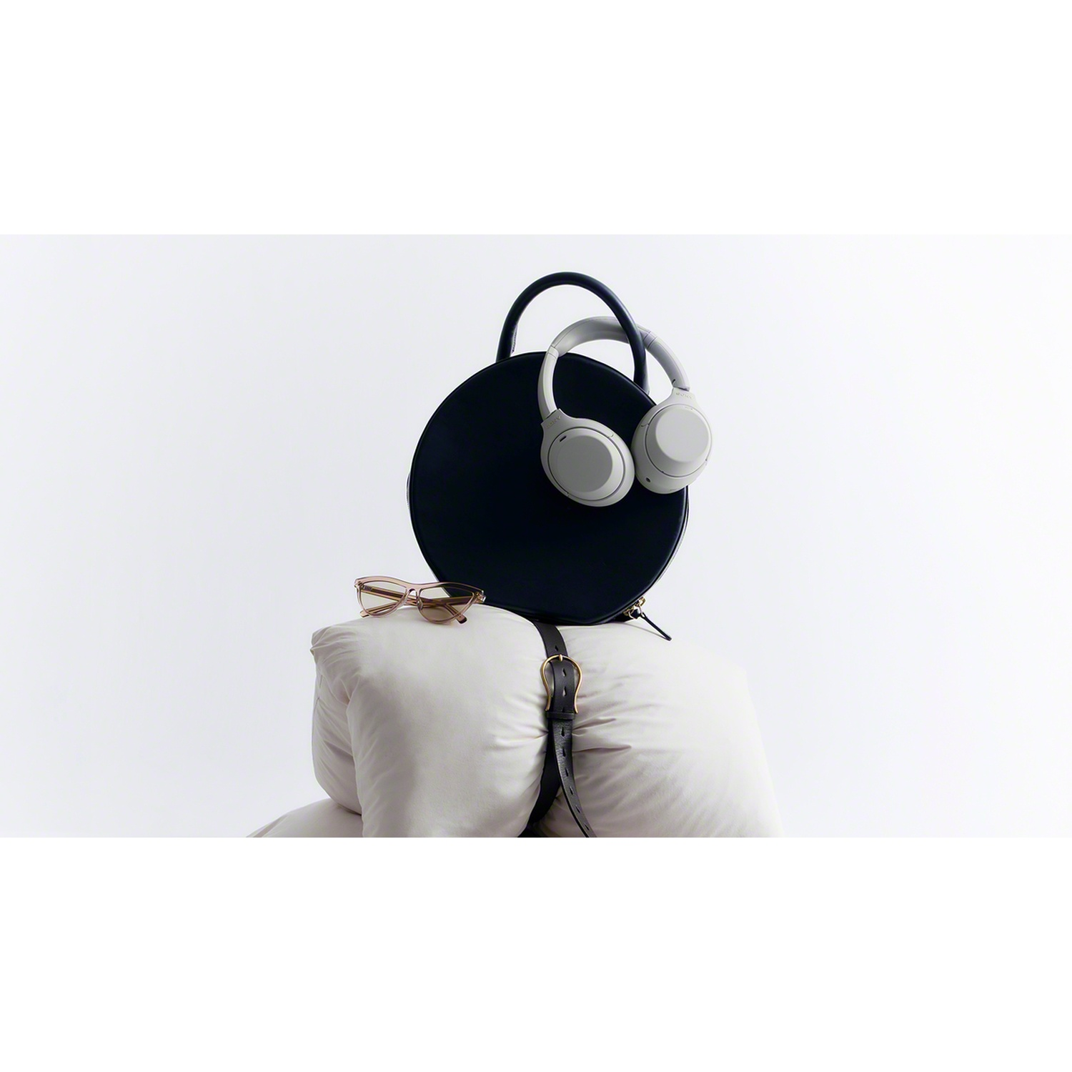 A round black handbag with headphones and glasses rests on white pillows against a white background.
