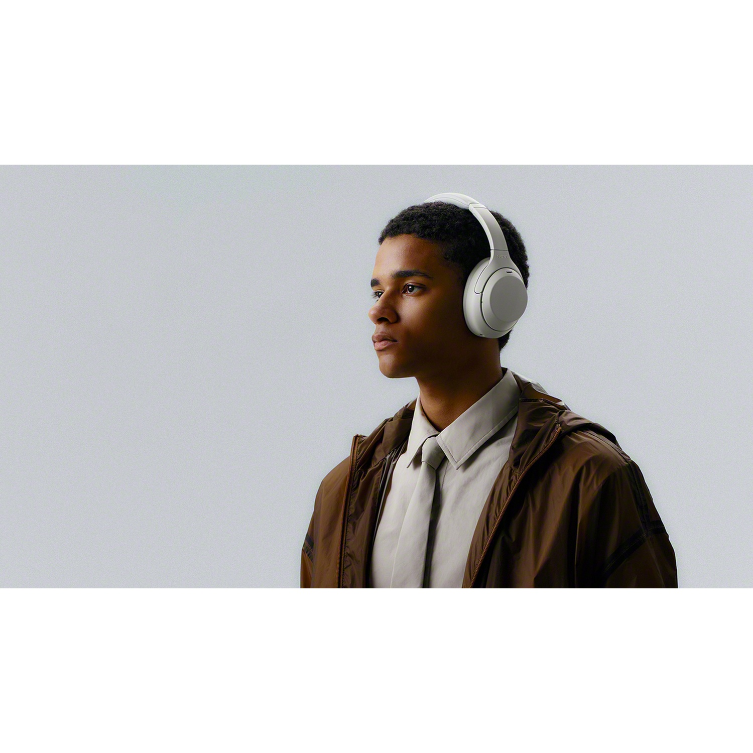 A young man wearing white headphones, a white shirt, and a brown jacket against a gray background.