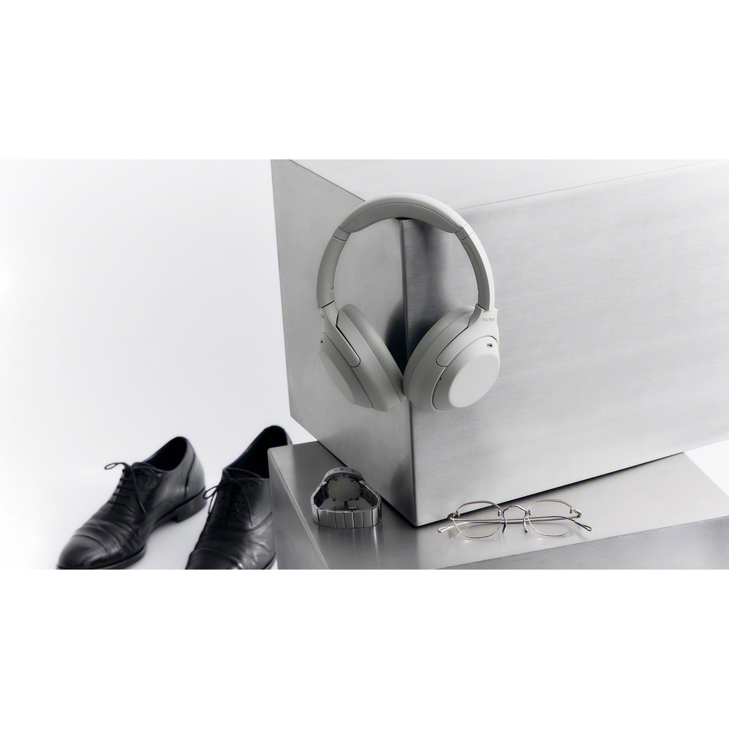 Grey headphones on a metal block, with a watch, glasses, and black shoes nearby.