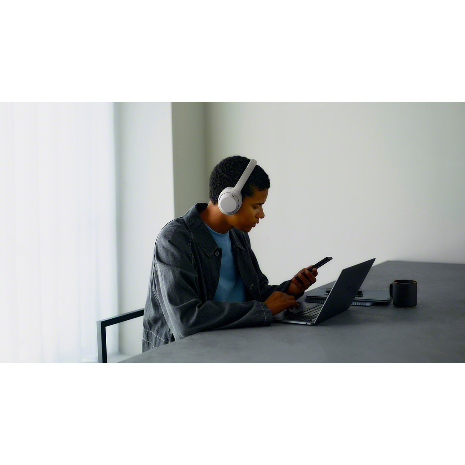 A man wearing headphones sits at a table, using a laptop and a smartphone.