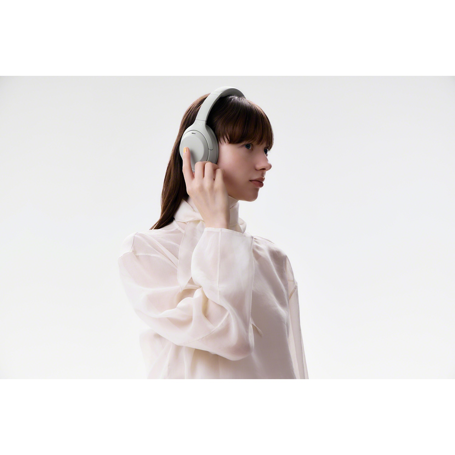 A woman wearing a white blouse touches her white over-ear headphones against a plain white background.