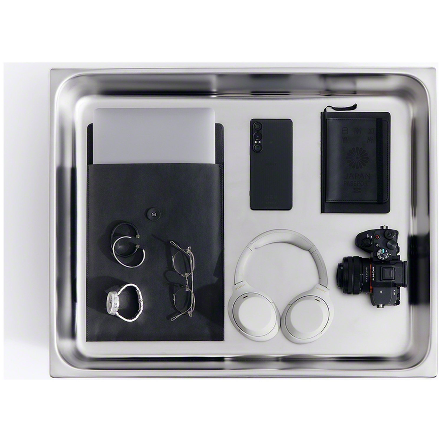 Flat stainless steel tray containing a laptop, smartphone, headphones, camera, glasses, watch, and Japan passport.