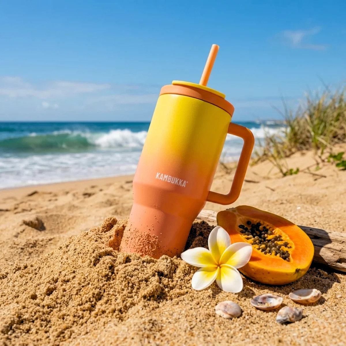 Pomarańczowo-żółty kubek ze słomką w piasku na plaży, obok papaja, kwiat i muszle.