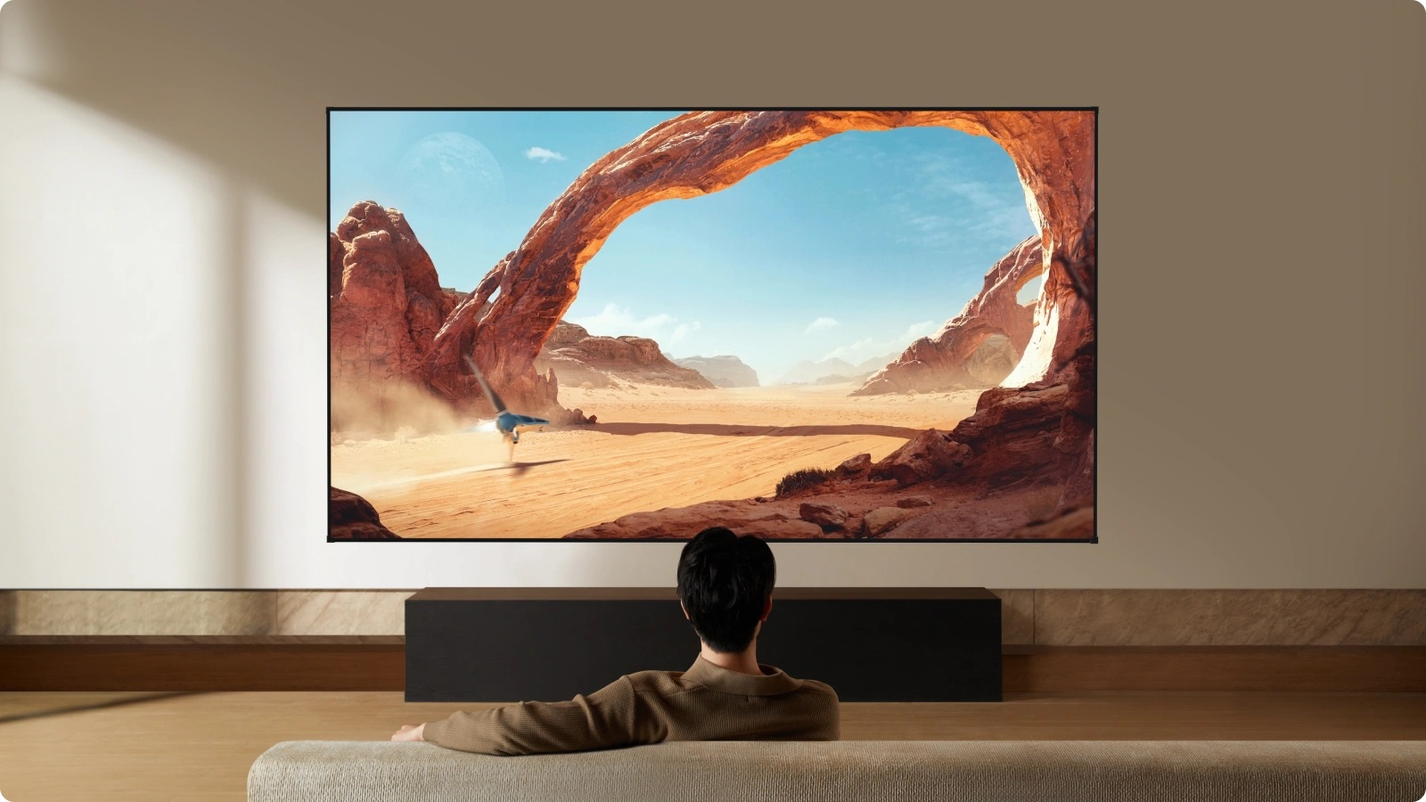 Bir adam, taş kemerli ve uçan cisimli çöl manzarası gösteren büyük bir TV'nin önünde oturuyor.