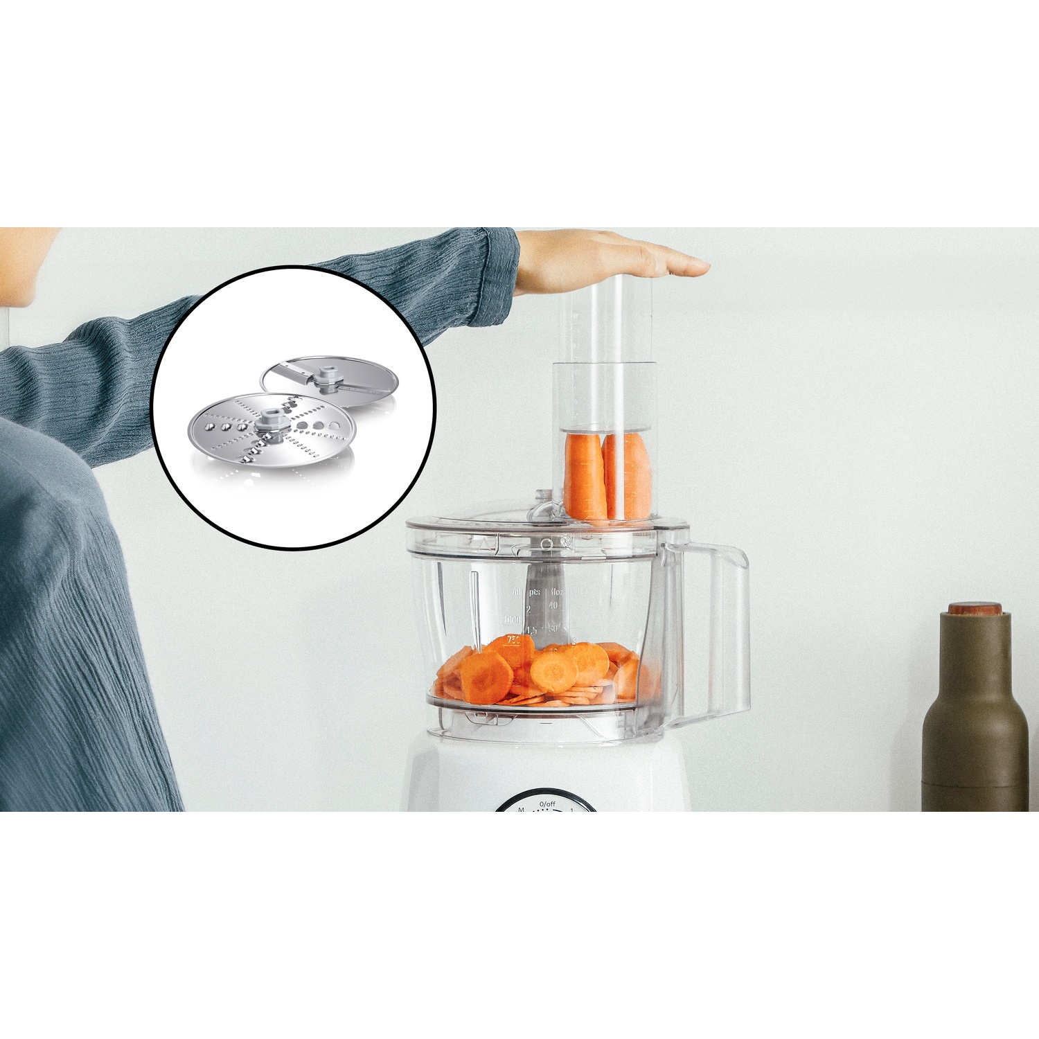 Una mano aziona un robot da cucina che affetta carote, con un riquadro che mostra le lame in acciaio.