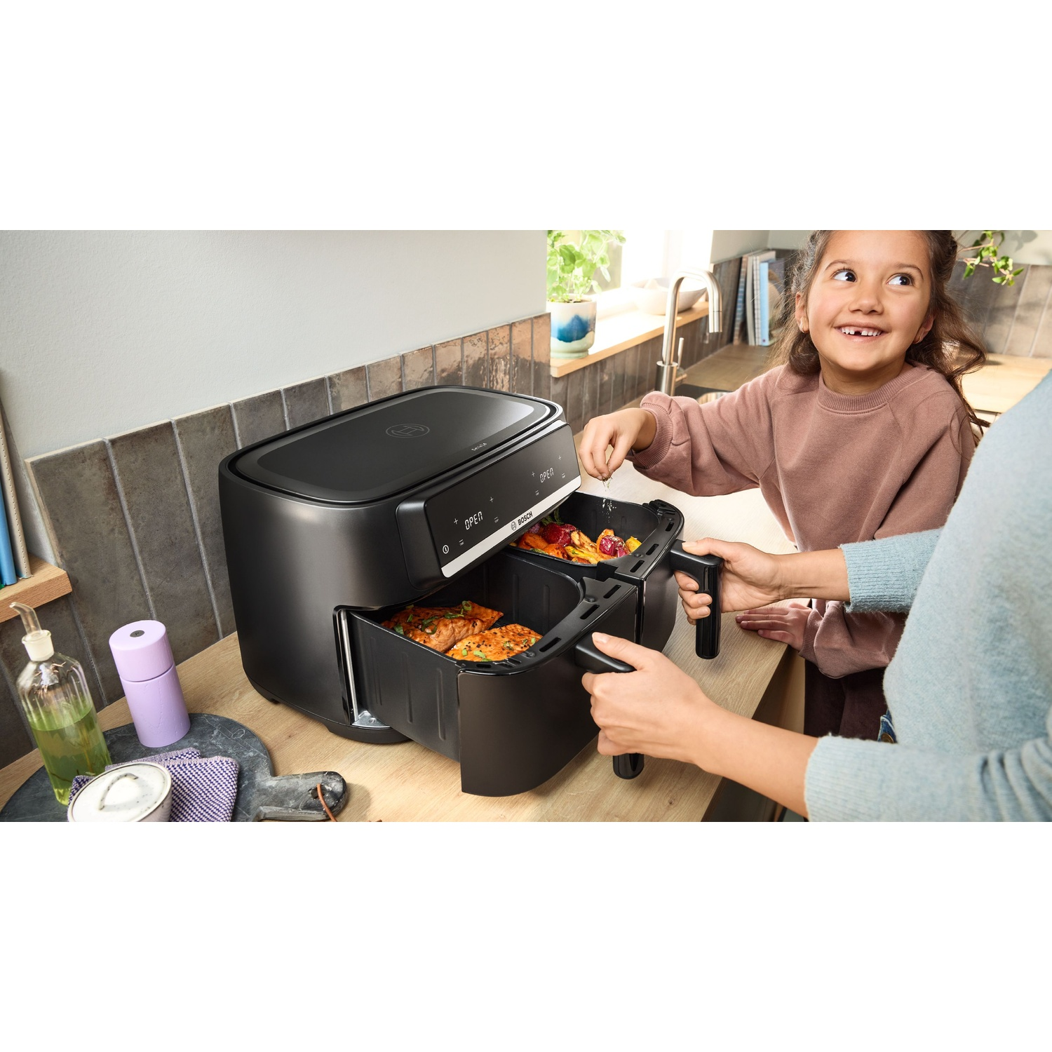 Una bambina aiuta ad aprire una friggitrice ad aria nera a doppio cassetto sul bancone della cucina.