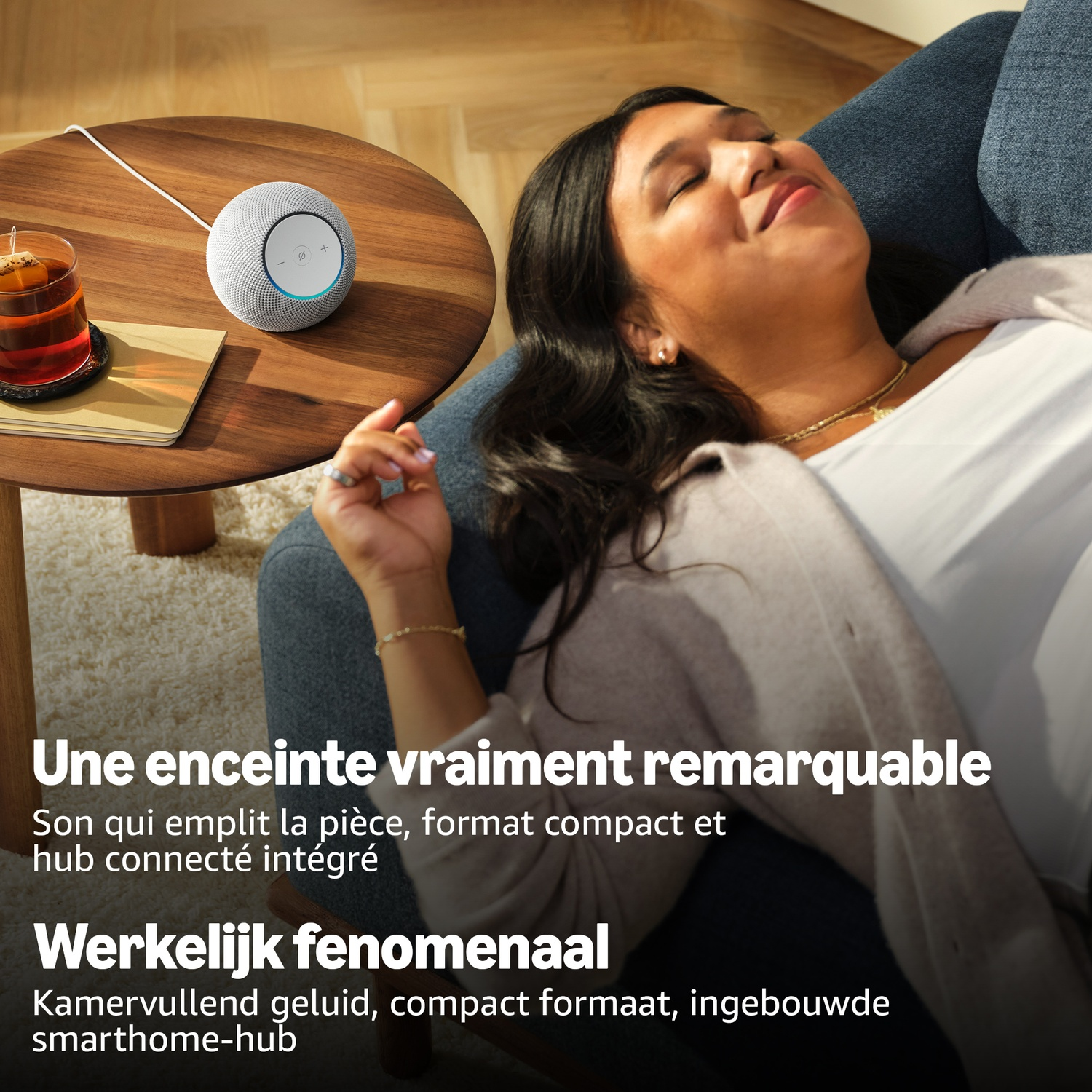 Une femme se détend sur un canapé bleu près d'une table en bois avec une enceinte connectée et du thé.