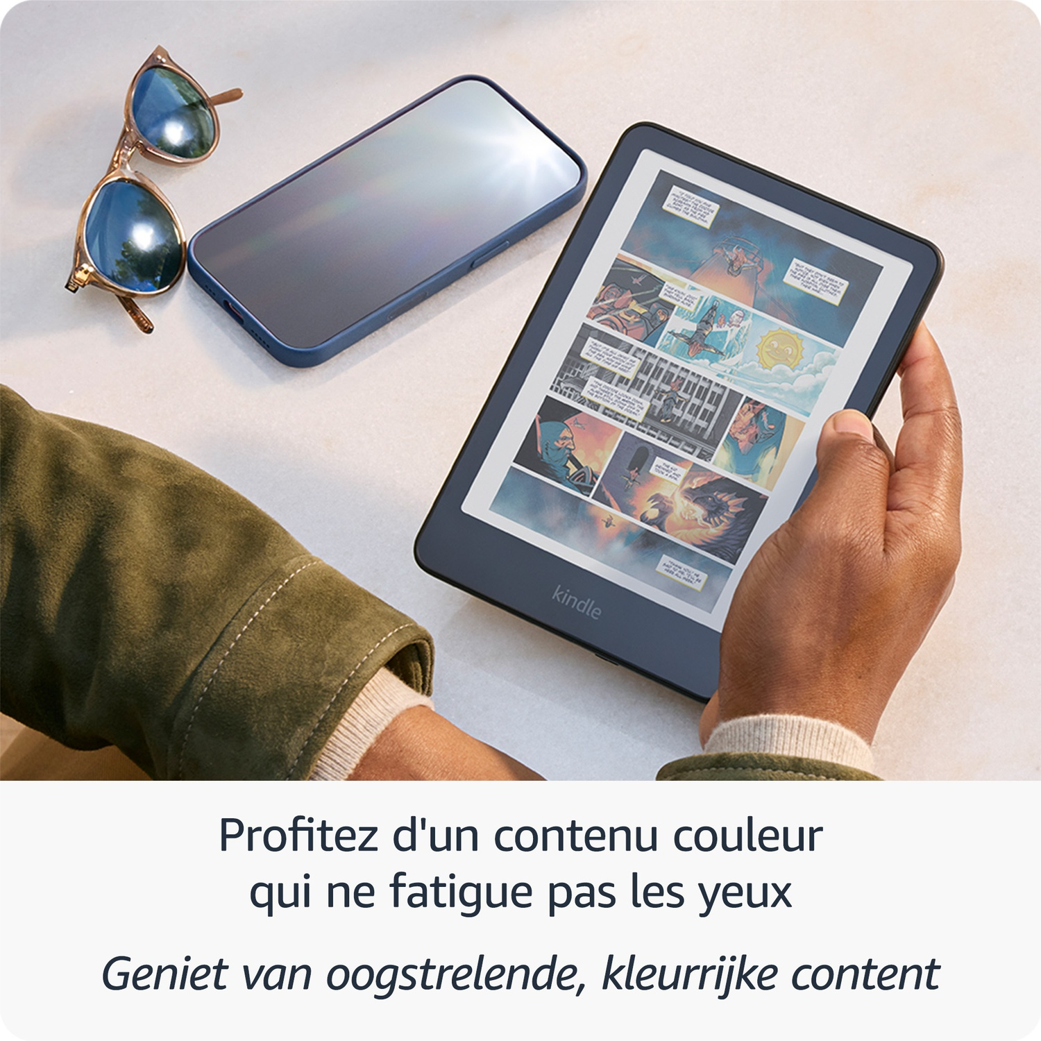Une personne tient une liseuse Kindle avec une BD en couleur; un téléphone et des lunettes sur une table.