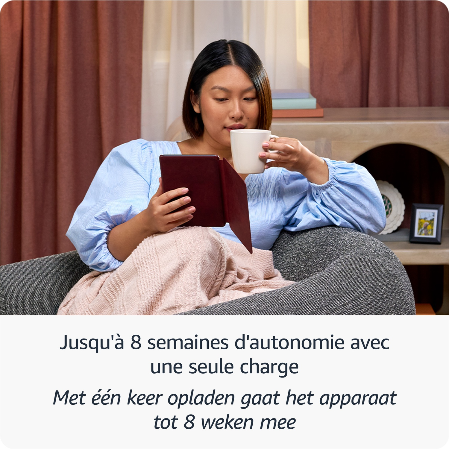 Une femme lit une liseuse dans un fauteuil gris, tenant une tasse blanche avec une couverture rose.