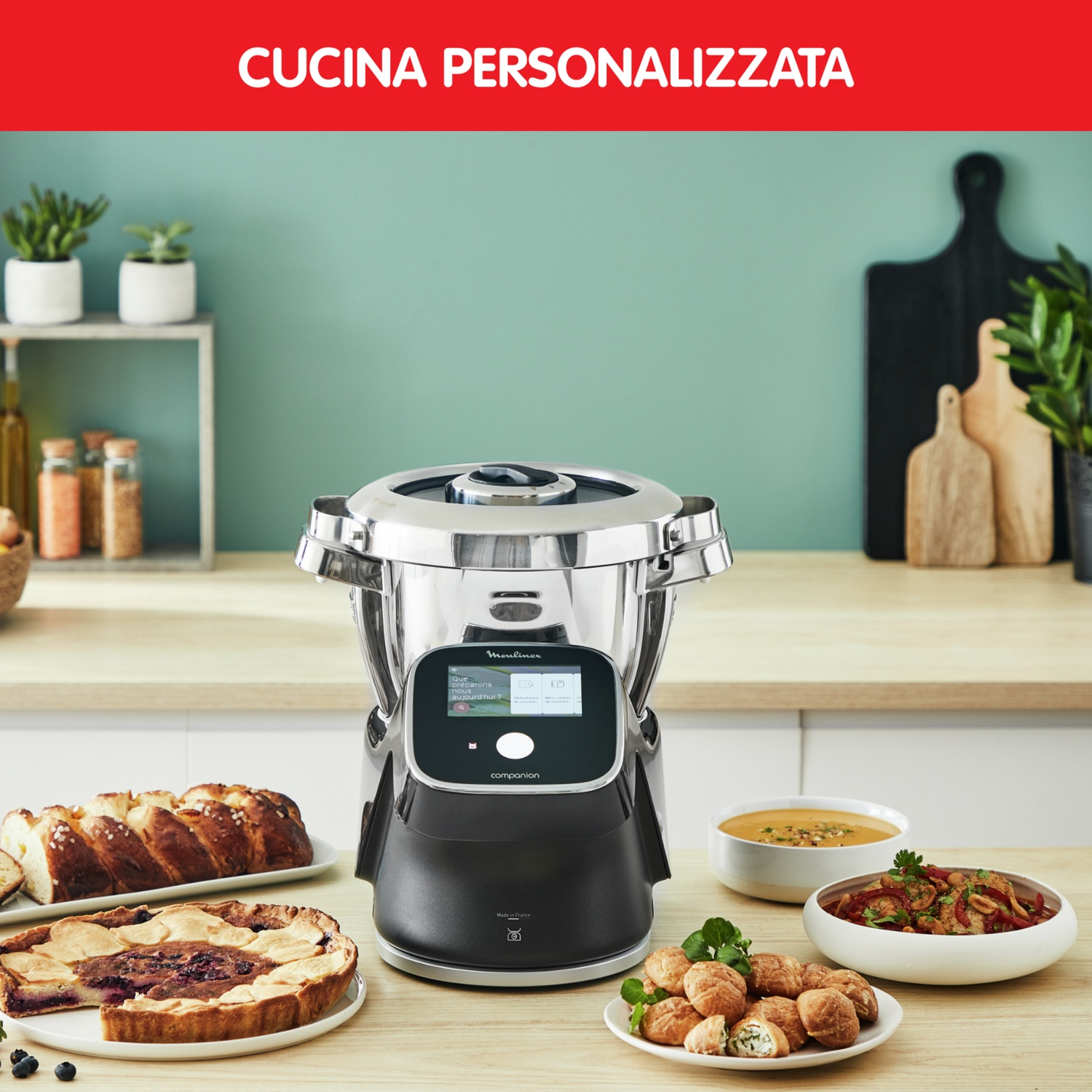 Un robot da cucina su un tavolo, circondato da pane, torta, zuppa e pasticcini in una cucina.