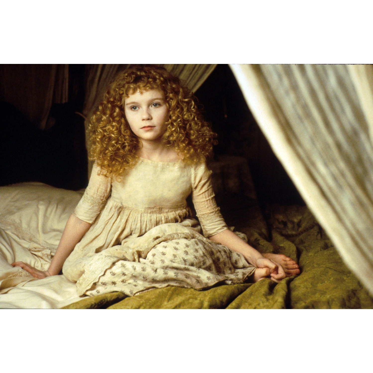 Une jeune fille aux cheveux bouclés blonds et robe beige assise sur un lit dans une pièce sombre.