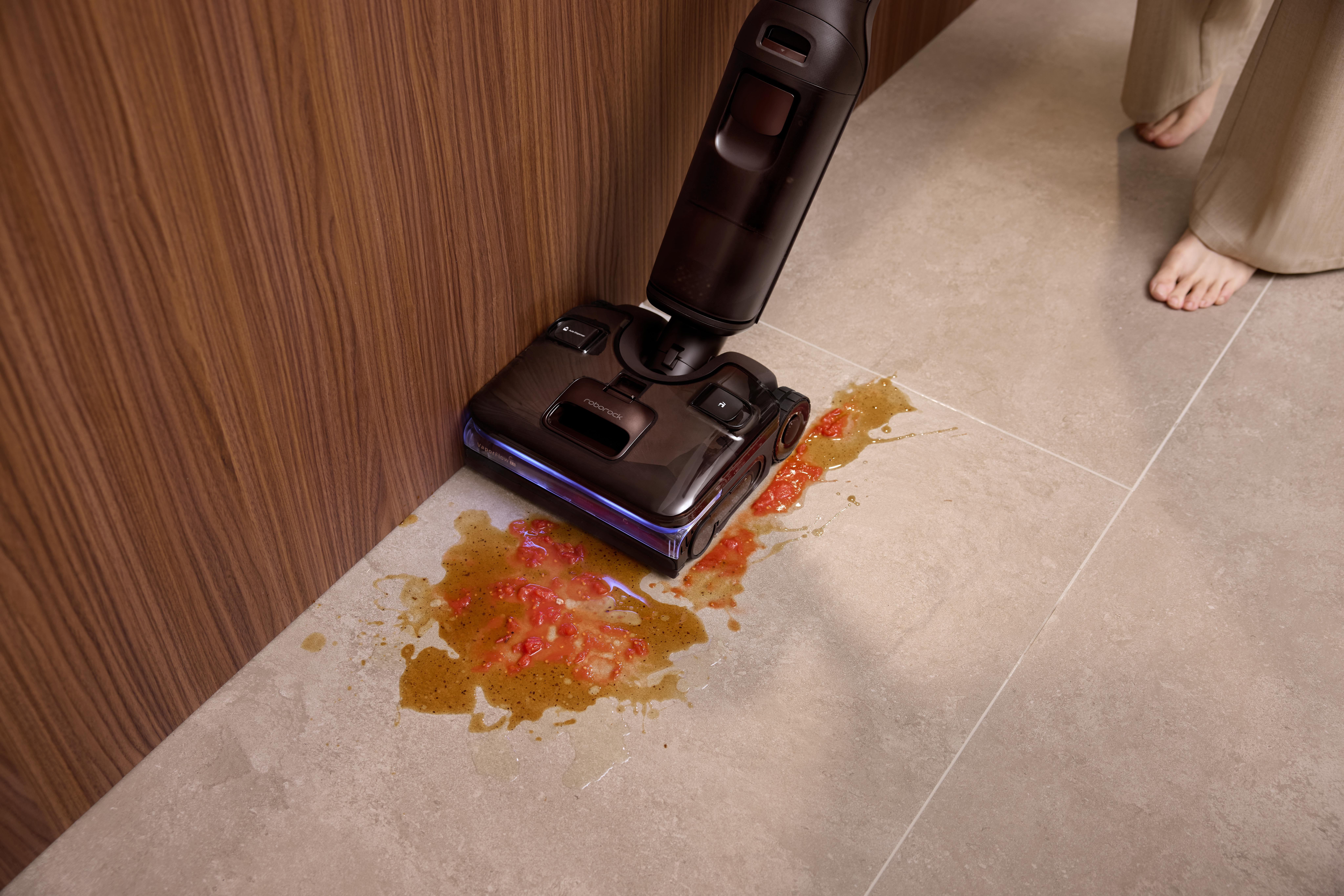 Ein schwarzer Nasssauger reinigt verschüttete Tomatensoße auf einem hellen Fliesenboden nahe einer holzverkleideten Wand.