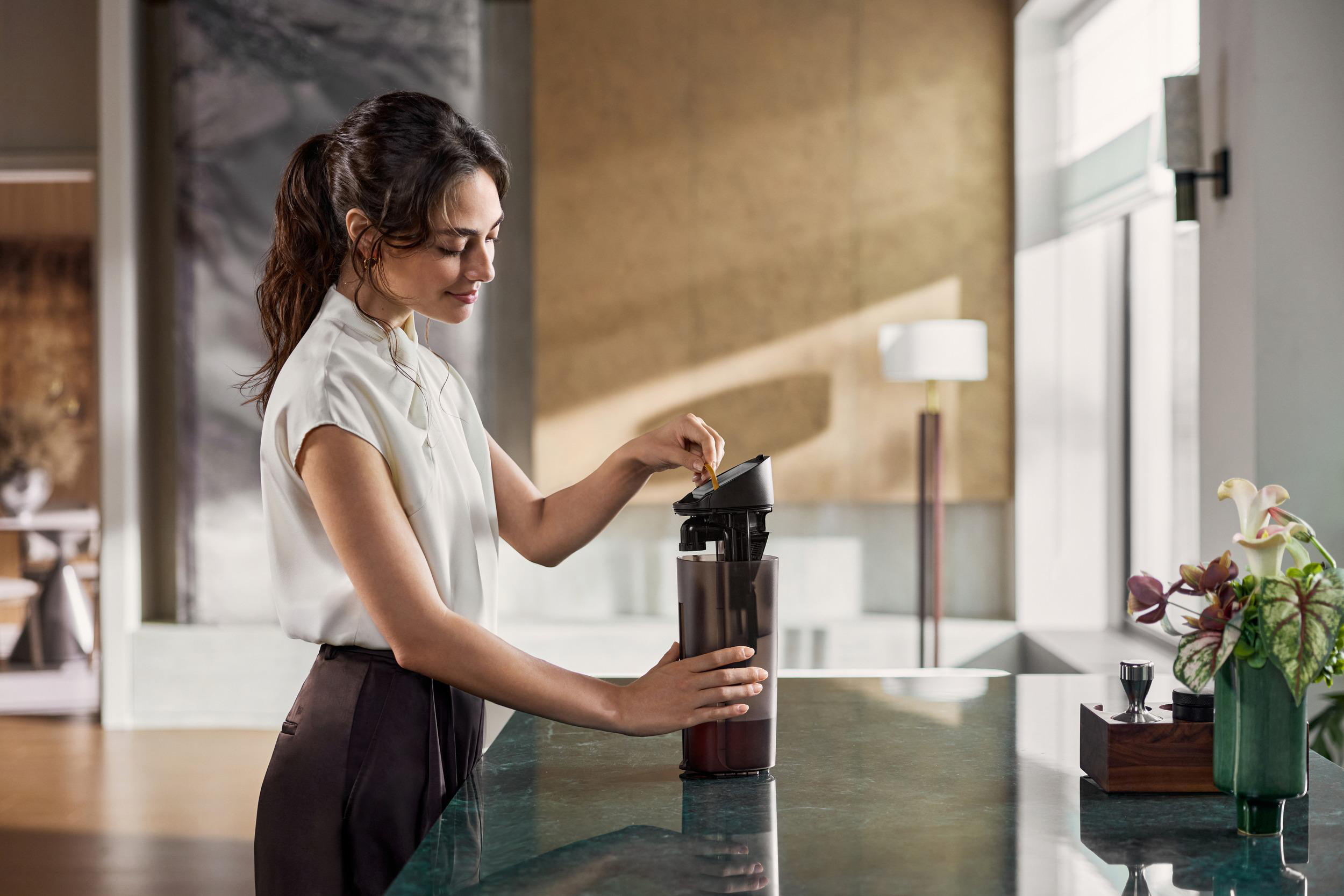 Eine Frau in weißer Bluse befüllt eine Kaffeemaschine in einer modernen Küche.