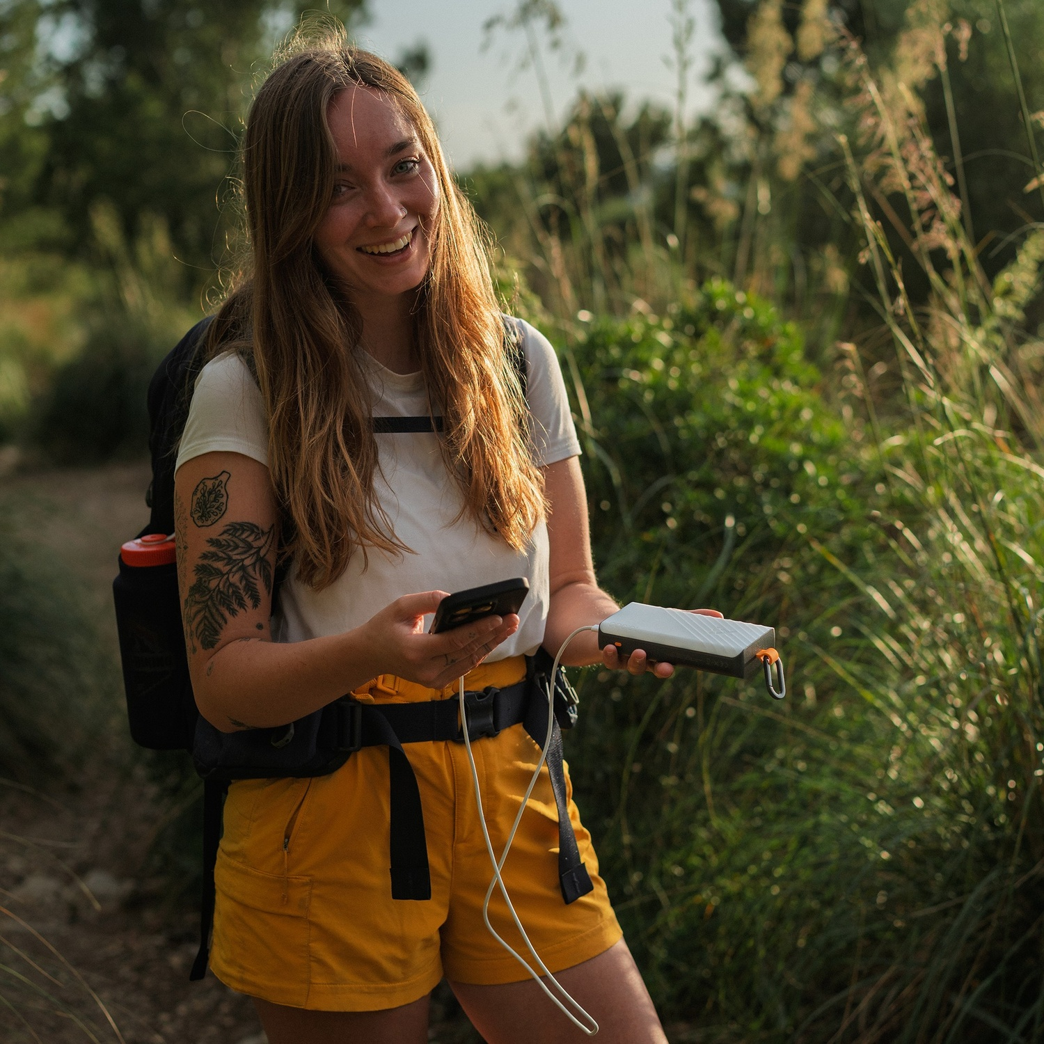 Randonneuse avec sac à dos sourit, tenant un smartphone et une batterie externe en plein air.