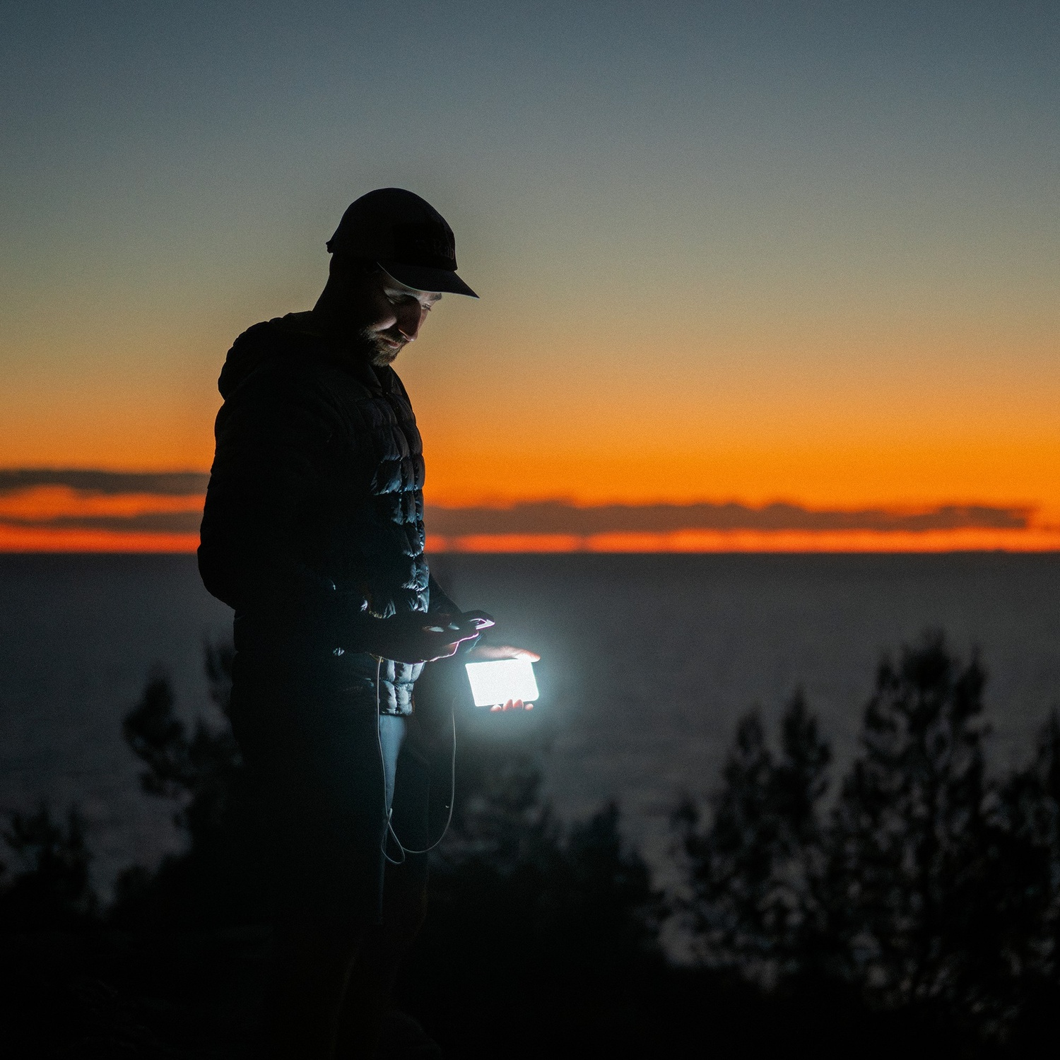 Un homme en sweat à capuche et casquette se tient près de la mer au crépuscule, tenant un smartphone lumineux.