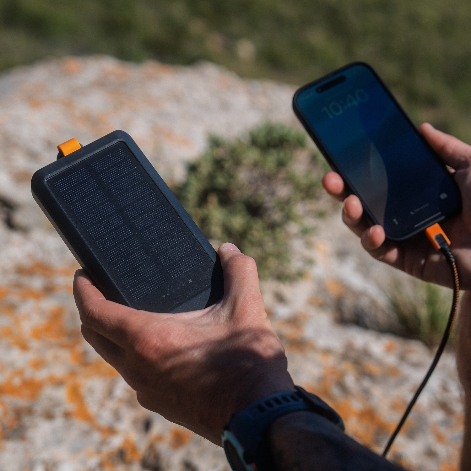 Une personne tient une batterie solaire noire et un smartphone avec un câble de charge à l'extérieur.