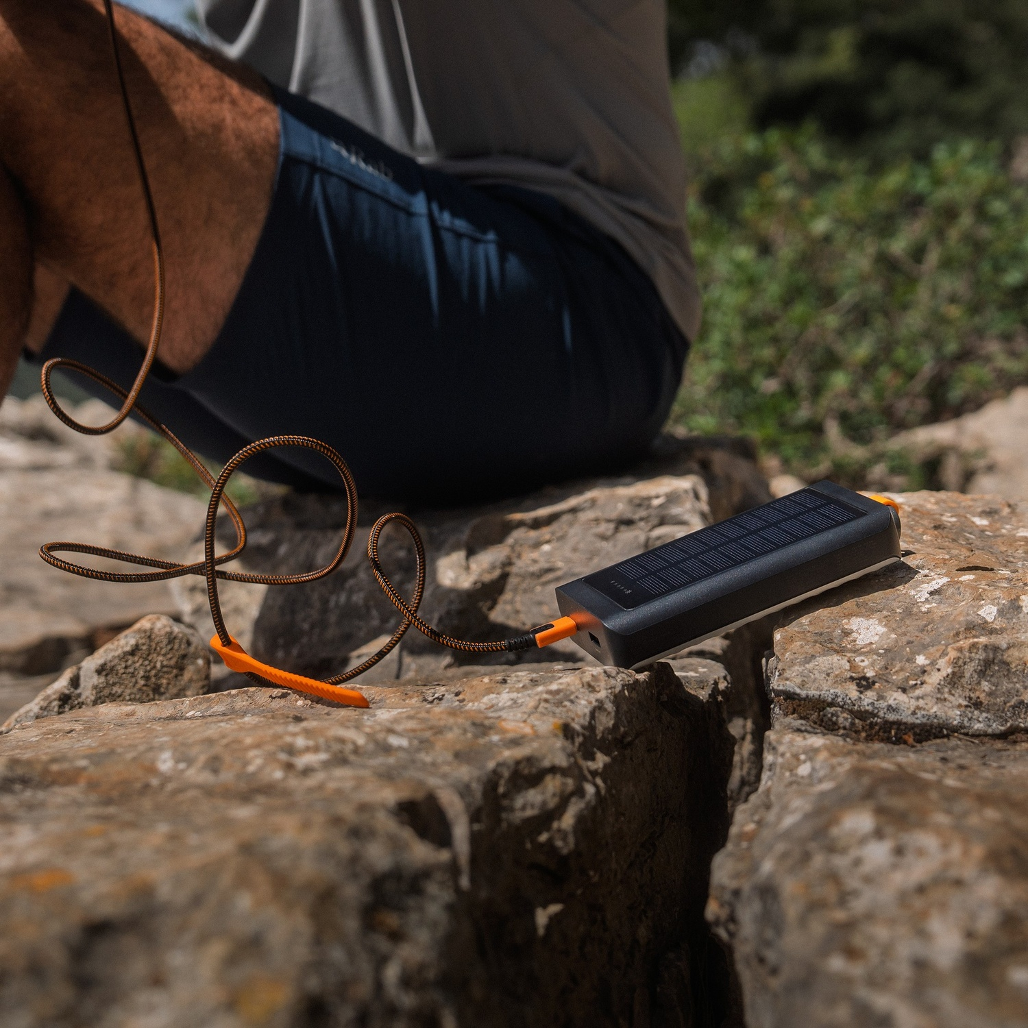 Une batterie externe avec câble repose sur un sol rocheux tandis qu'une personne est assise sur des pierres.