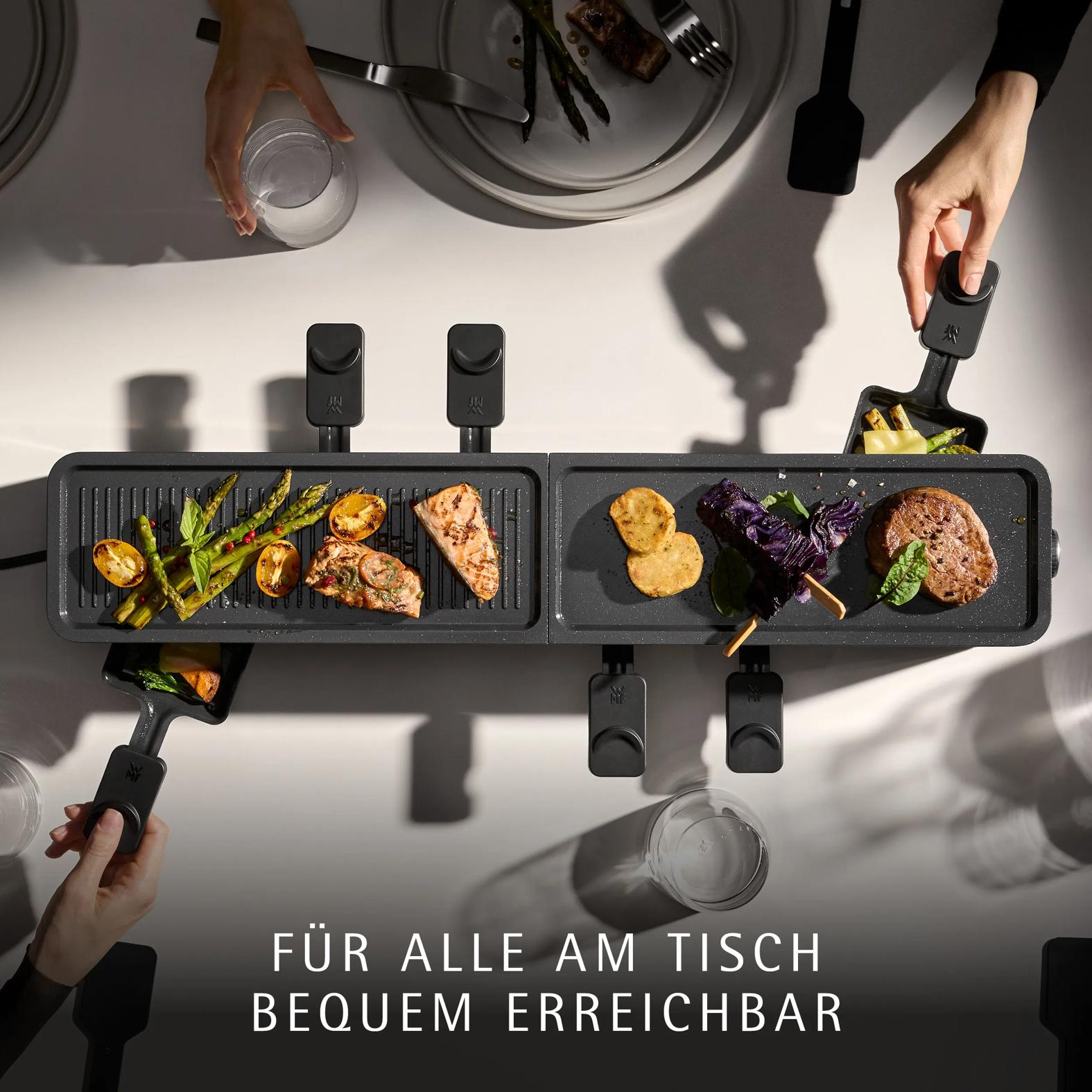 Top-down view of people using a long black raclette grill with asparagus, fish, vegetables, and meat.