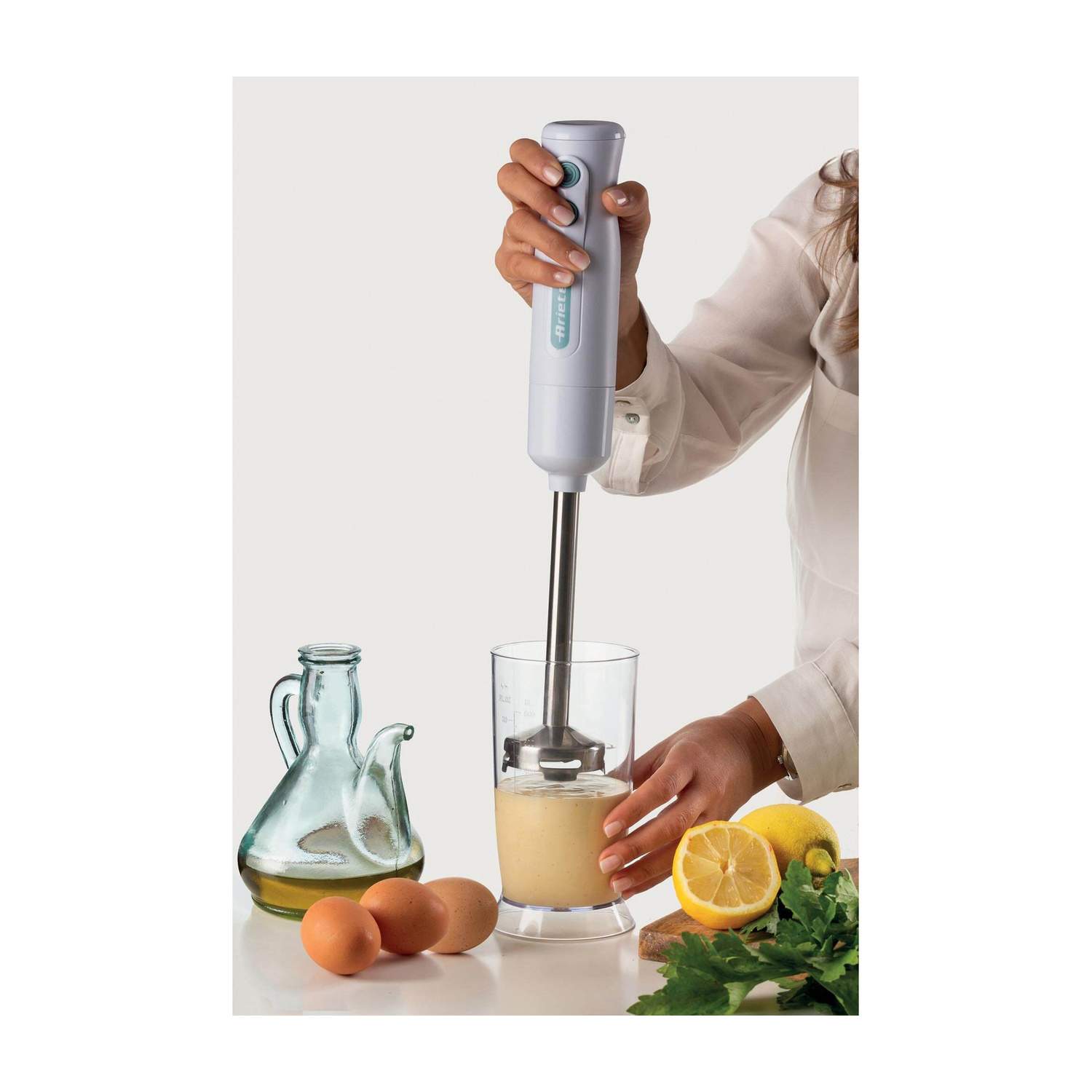 Una donna tiene un frullatore a immersione in un bicchiere di liquido, circondata da olio, uova e limoni.