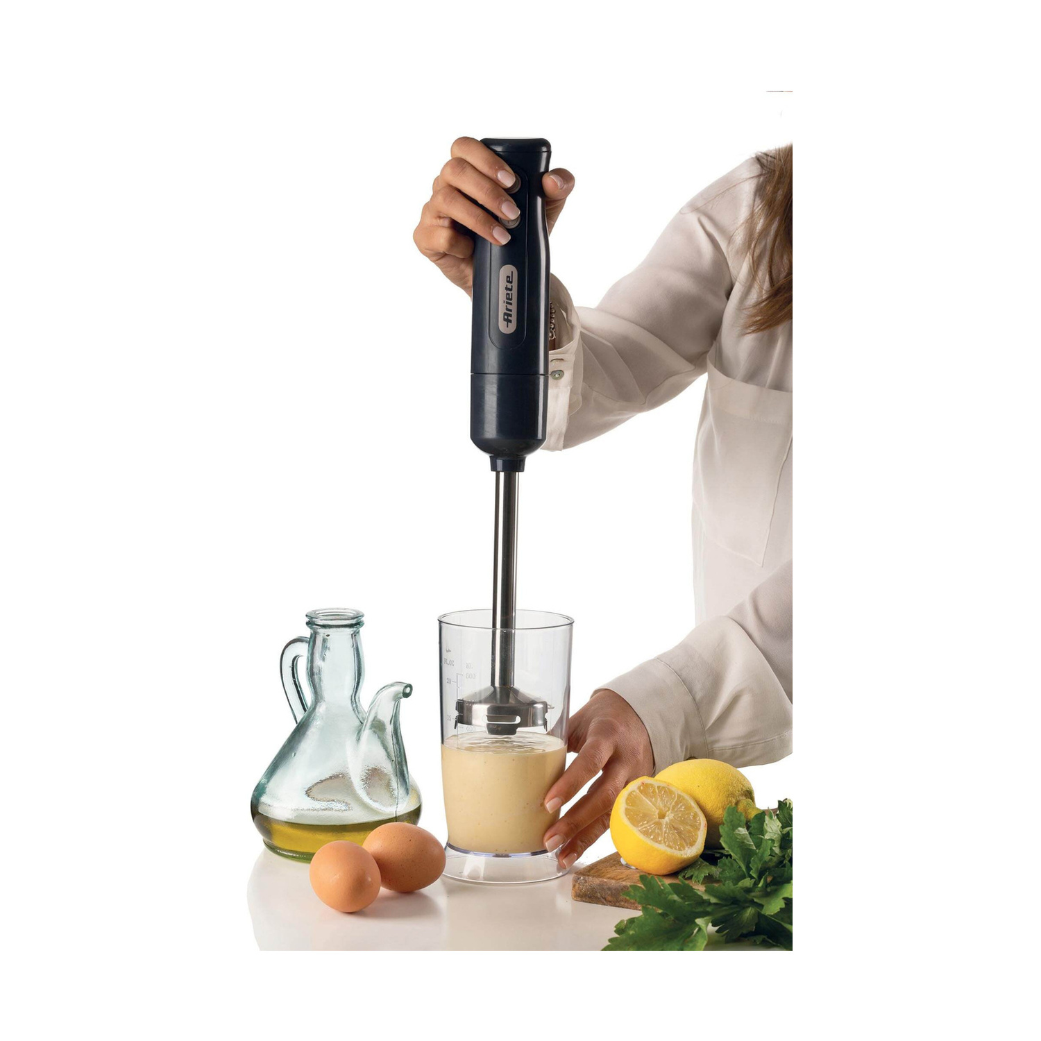 Una persona usa un frullatore ad immersione in un bicchiere di liquido, con olio, uova e limoni vicini.