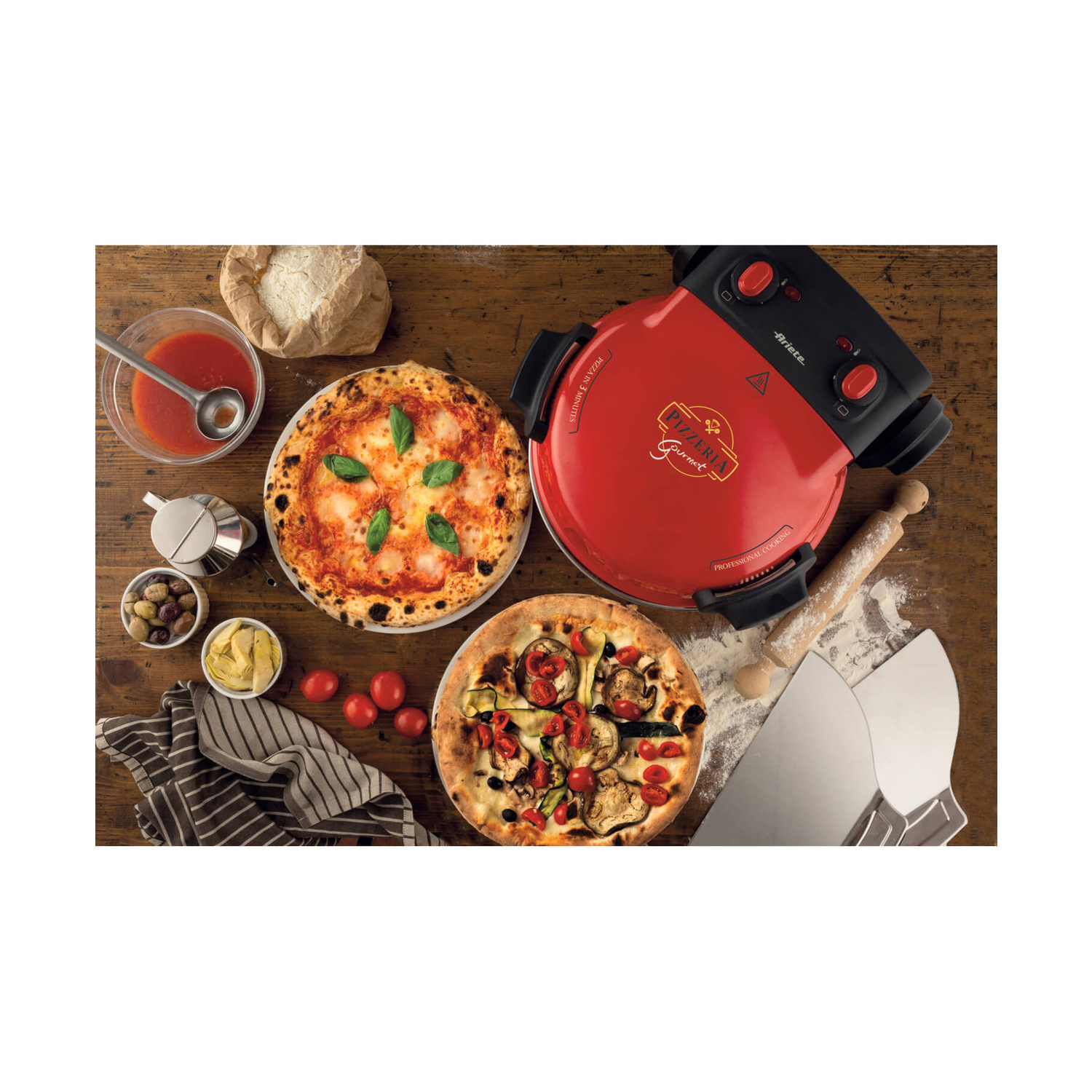 Forno per pizza rosso su tavolo in legno con due pizze, ingredienti e mattarello.