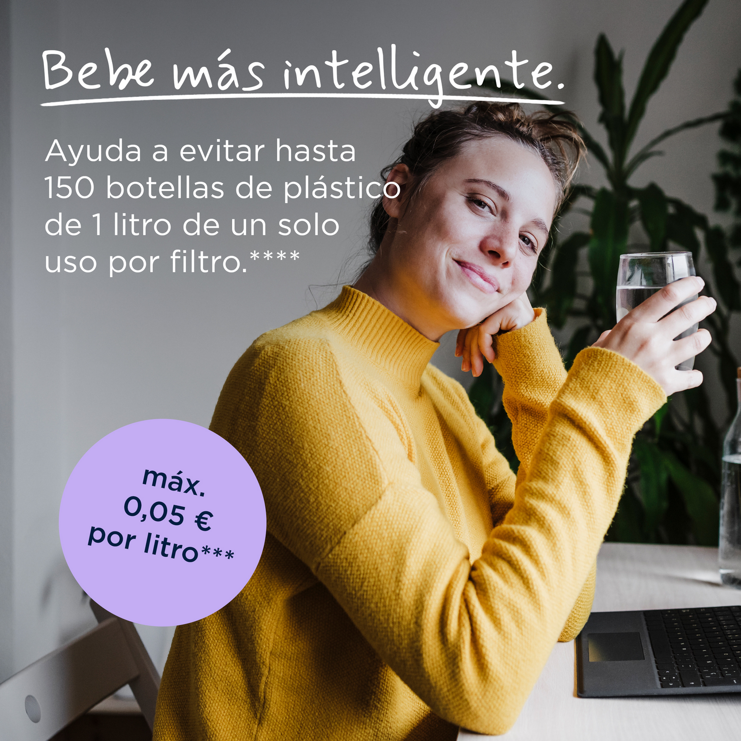 Una mujer con suéter amarillo está sentada con un vaso de agua en un portátil, plantas al fondo.