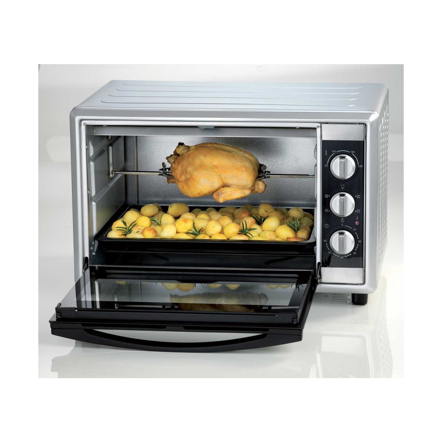 Un mini forno argento con pollo arrosto su uno spiedo sopra le patate.