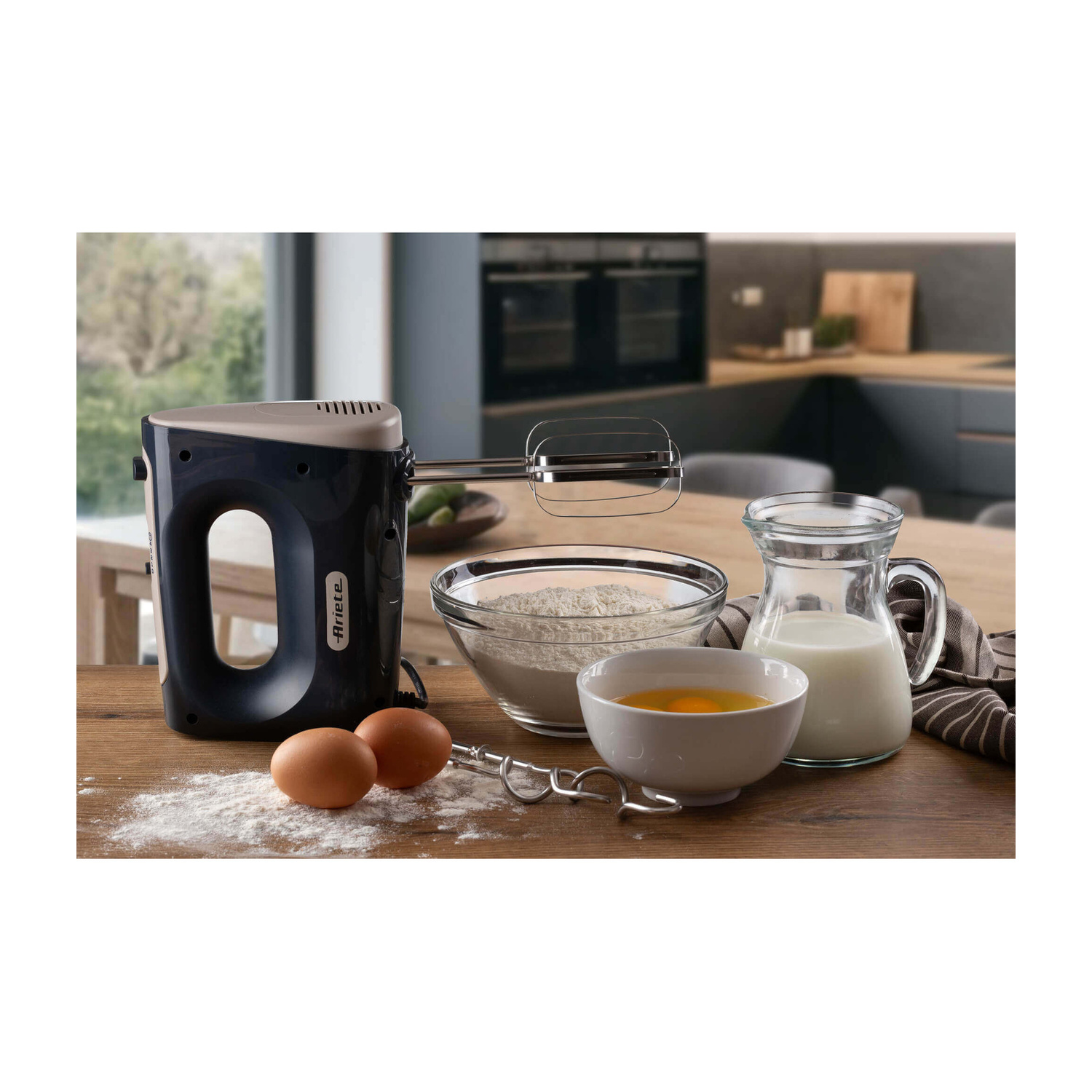 Un mixer a mano blu su un tavolo di legno con farina, uova e una brocca di latte in una cucina.