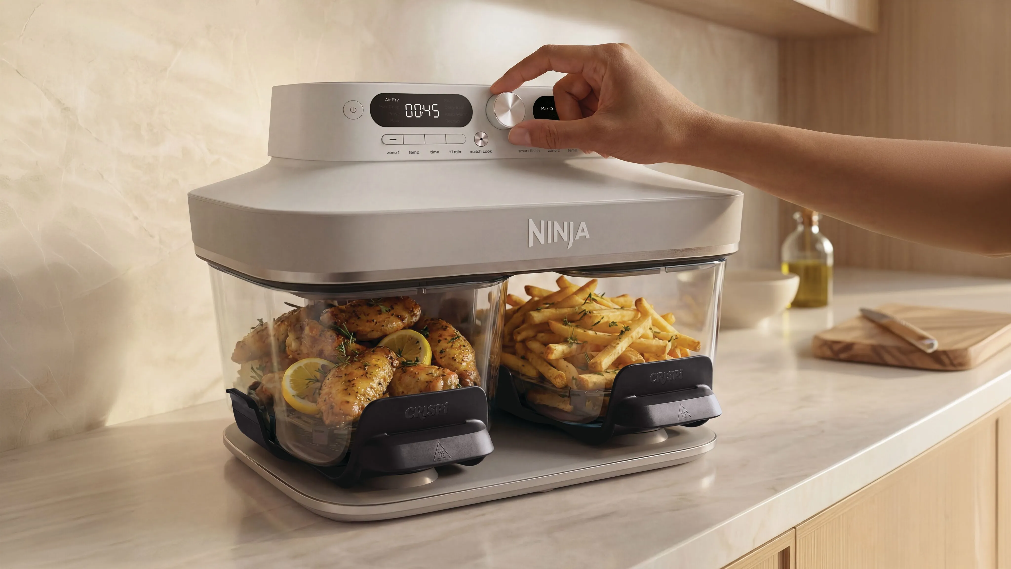 Hand bedient eine weiße Ninja Heißluftfritteuse mit zwei durchsichtigen Schubladen für Hähnchen und Pommes.