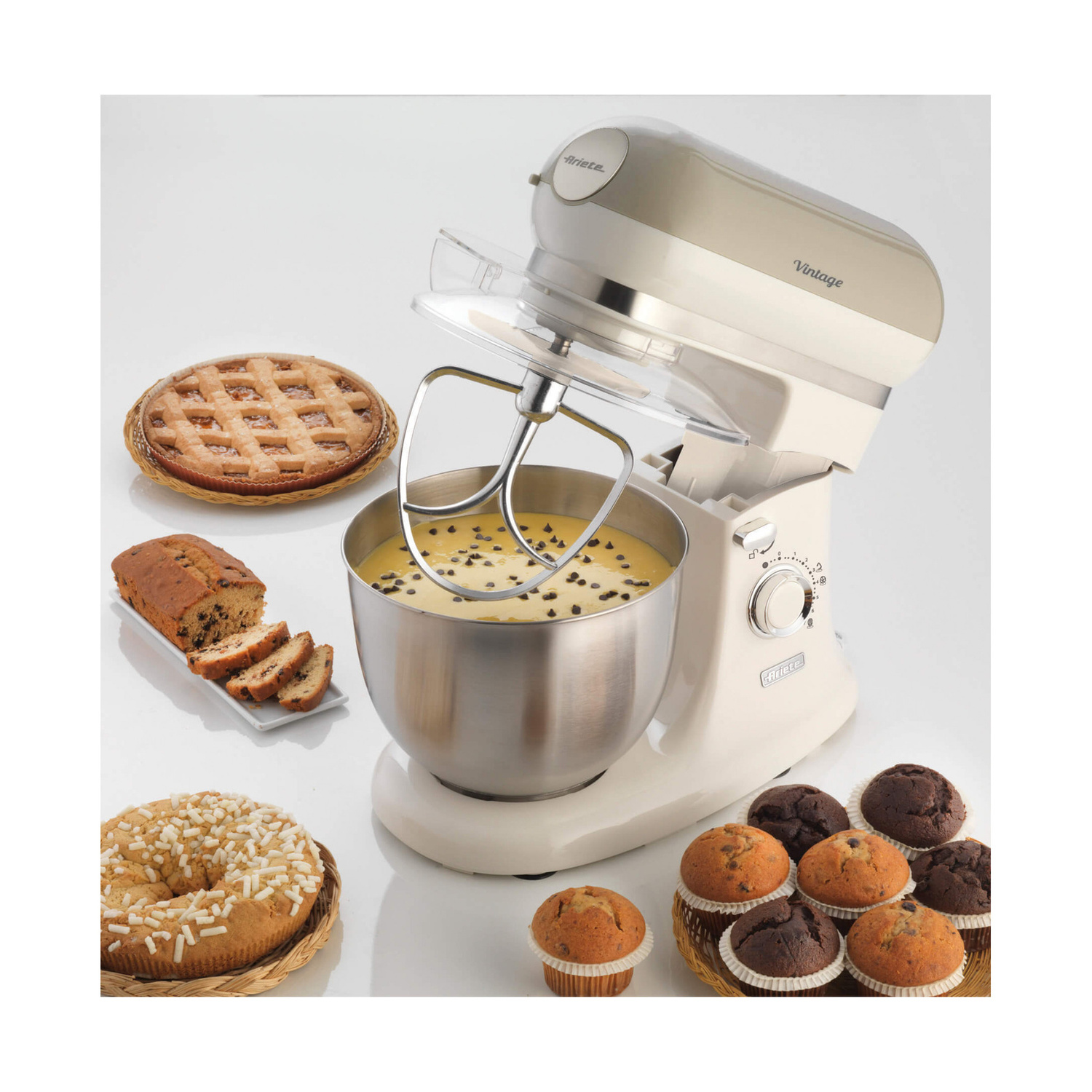 Un'impastatrice color crema con ciotola di impasto, circondata da torte cotte, muffin e una crostata su un tavolo bianco.