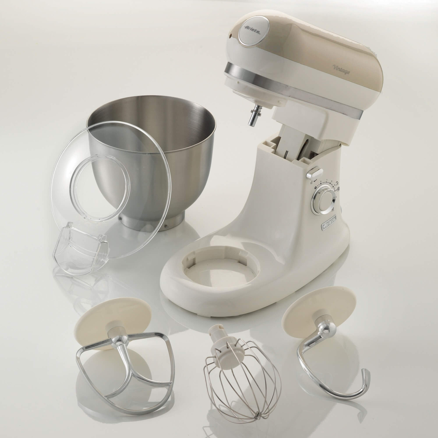 Impastatrice beige con ciotola in acciaio inox, coperchio trasparente e tre accessori su superficie bianca.