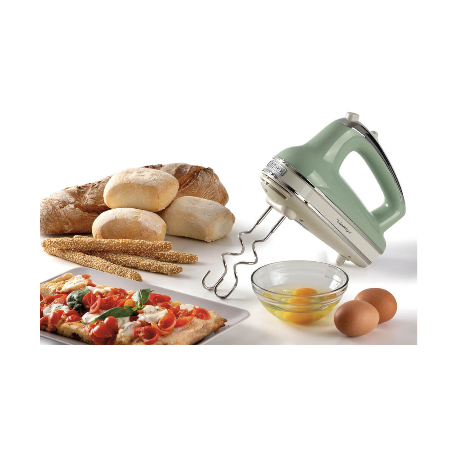 Un mixer a mano verde, pane, pizza, uova e una ciotola con tuorli su sfondo bianco.