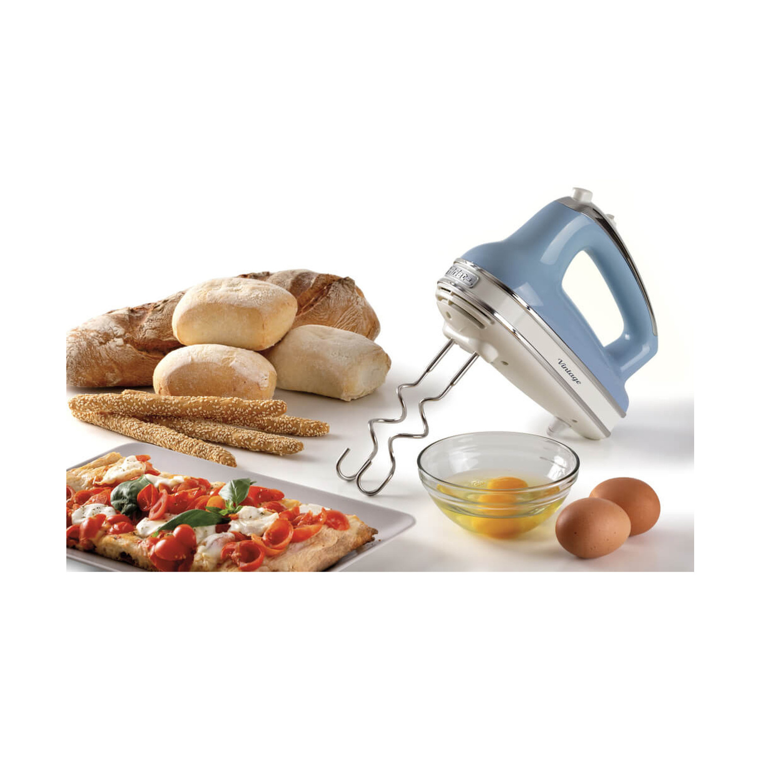 Un sbattitore azzurro con ganci, ciotola con uova, uova intere, pane, grissini e una focaccia al pomodoro.