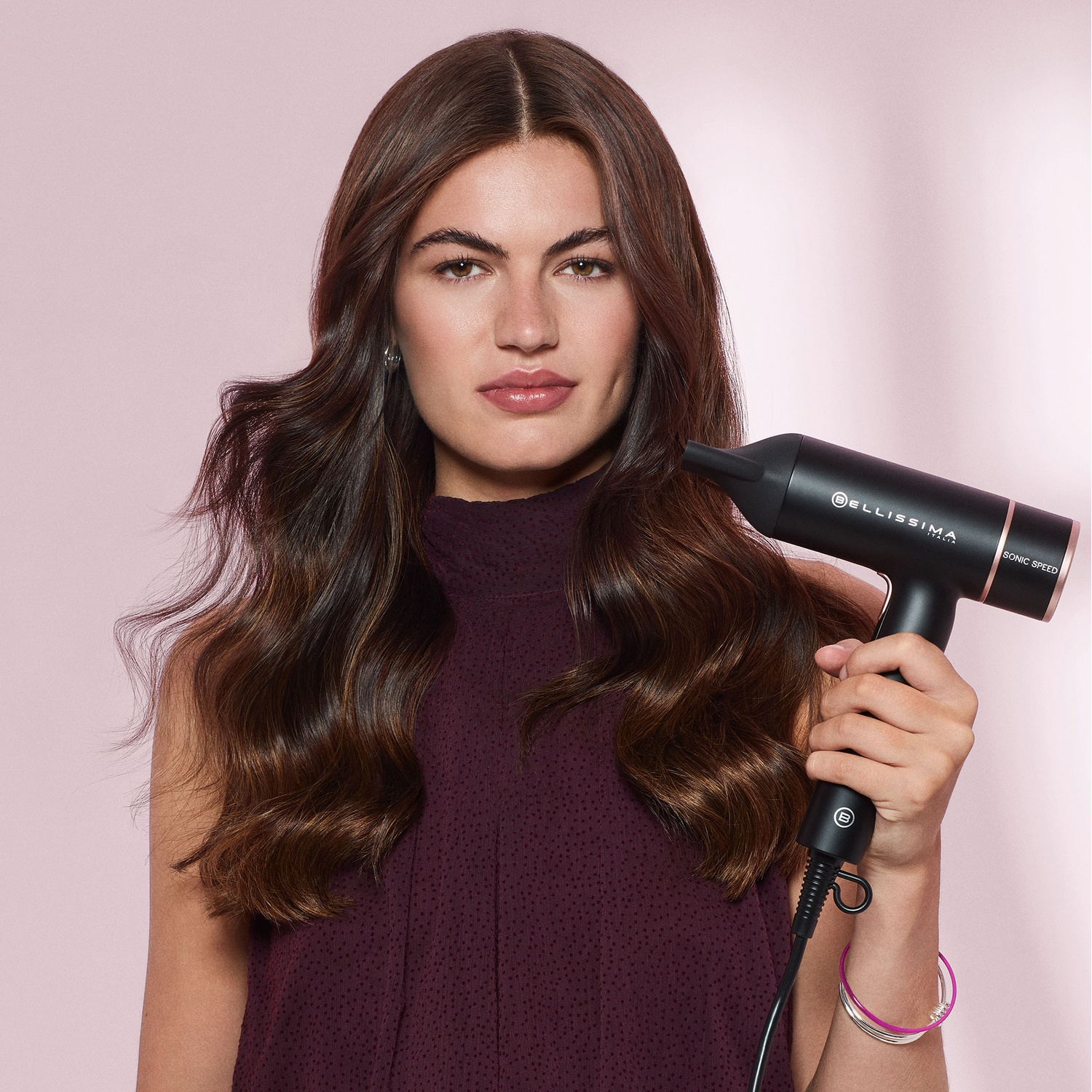 Una donna dai capelli castani tiene in mano un asciugacapelli Bellissima nero davanti a uno sfondo rosa.
