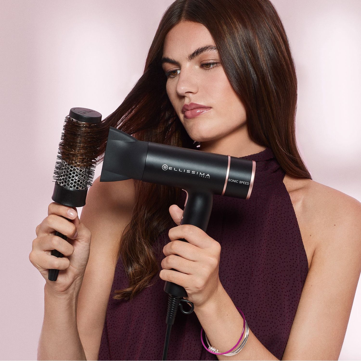 Una donna con lunghi capelli castani asciuga una ciocca con phon e spazzola tonda, sfondo rosa.