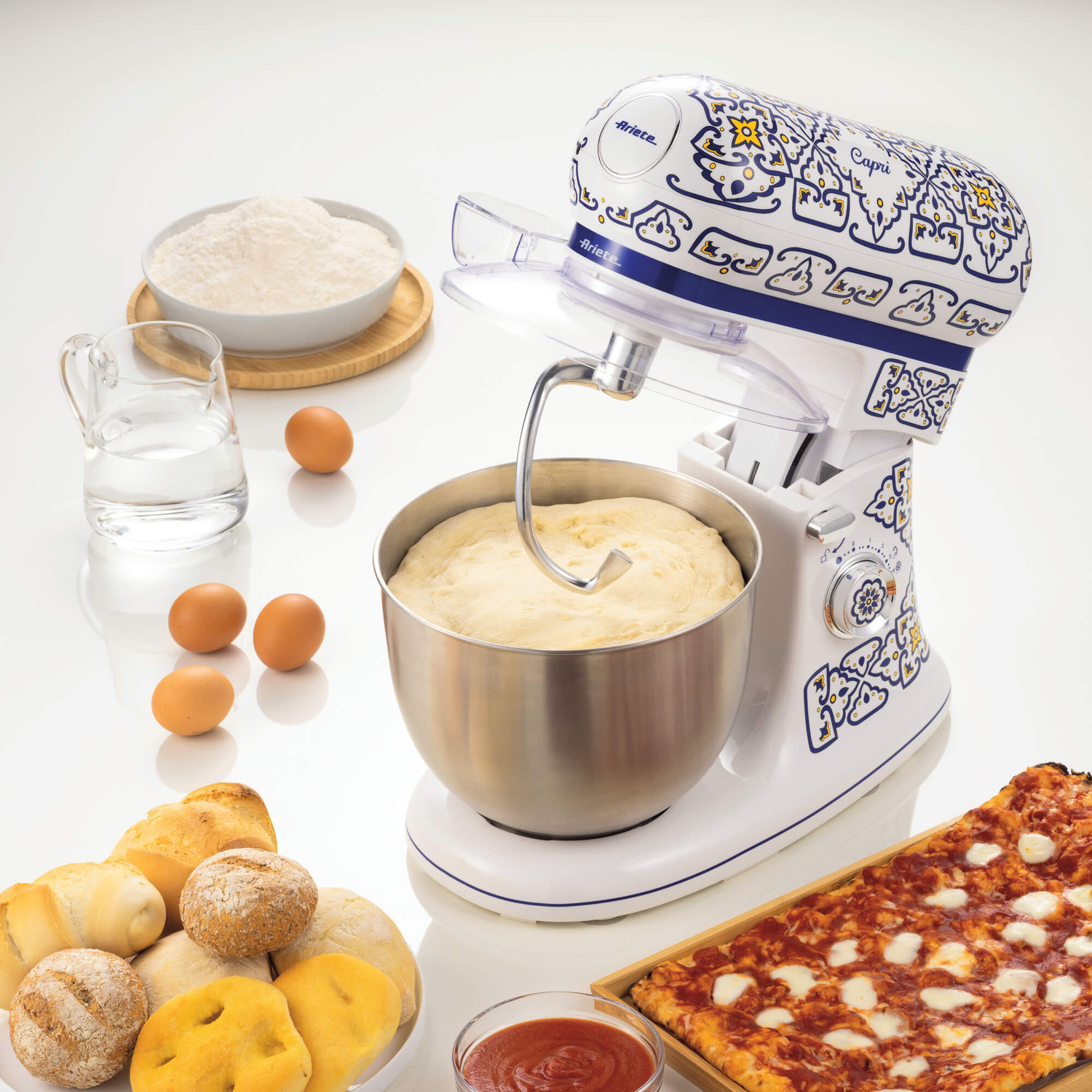 Impastatrice bianco-blu con ciotola, farina, uova, acqua e pizza su sfondo bianco.