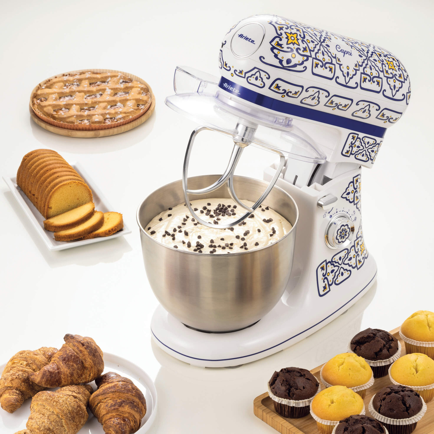 Un'impastatrice bianca con motivi mediterranei si trova accanto a prodotti da forno su un tavolo bianco.