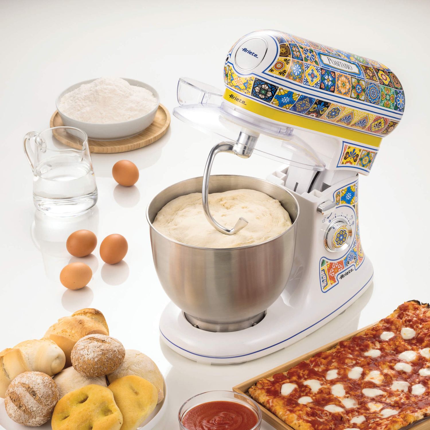 Impastatrice bianca con motivi colorati, ciotola, farina, acqua, uova, pane e pizza su tavolo bianco.