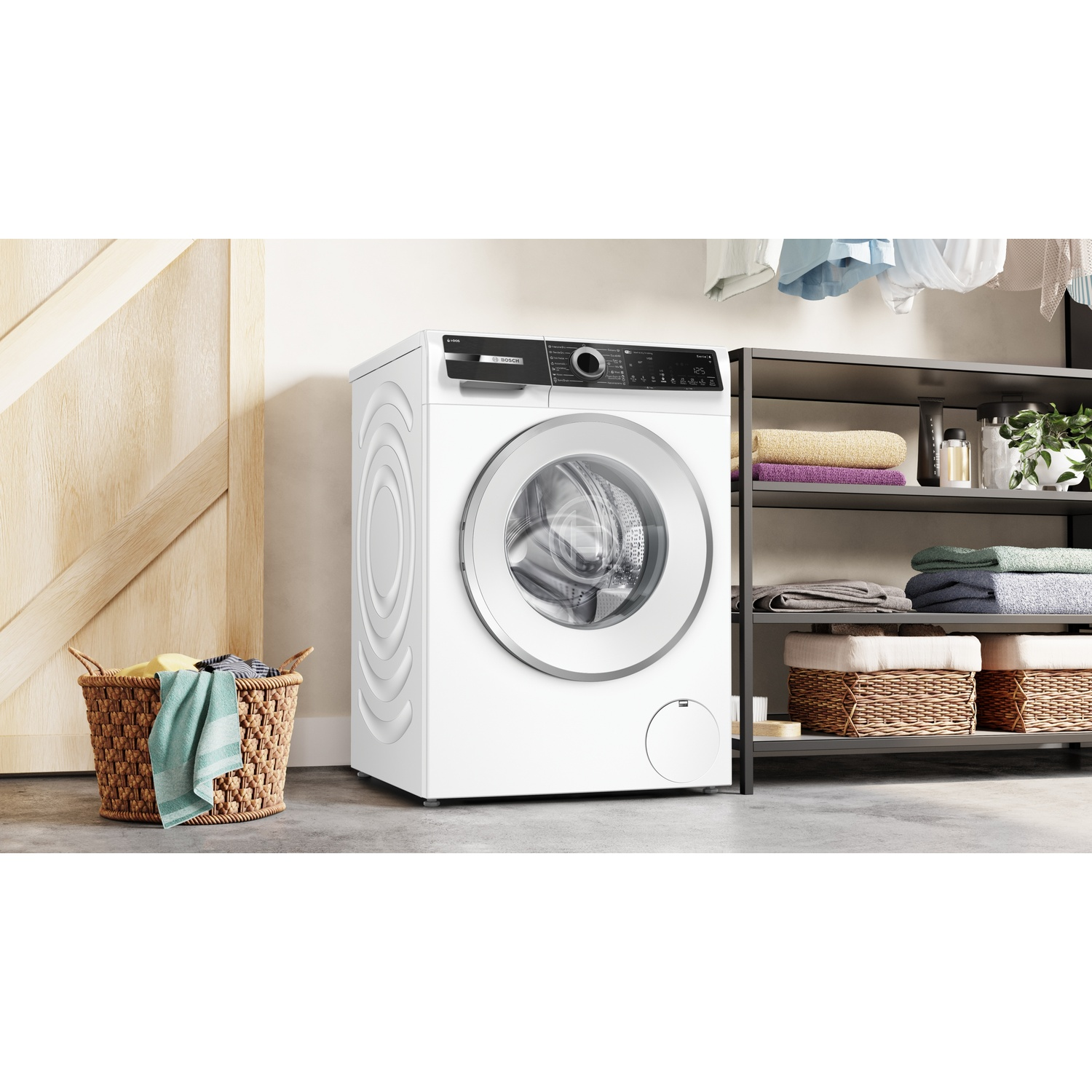 Een witte wasmachine staat in een lichte kamer naast een mand met wasgoed en een plank.